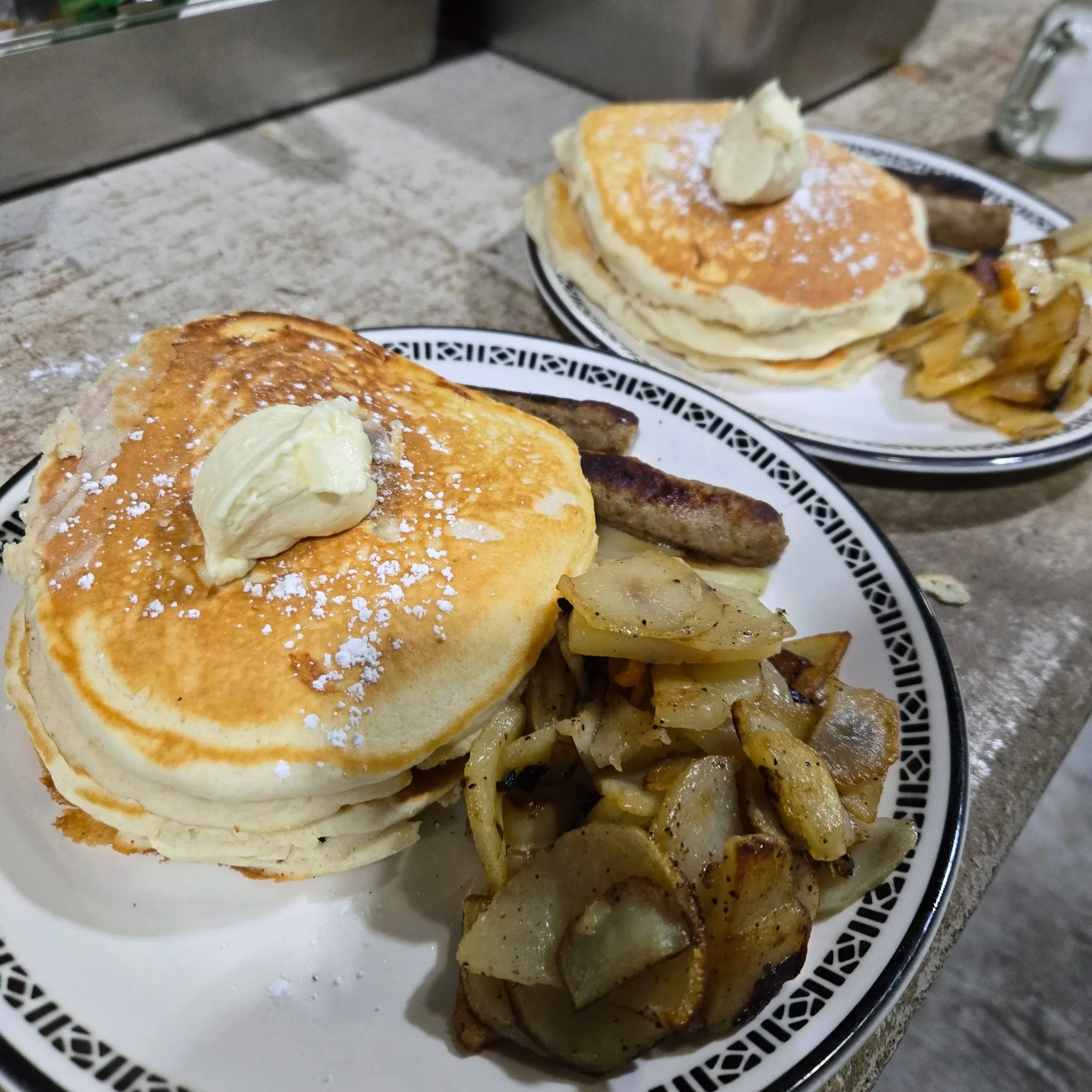 Two plates of pancakes topped with butter and powdered sugar, served with breakfast sausage and roasted potatoes.