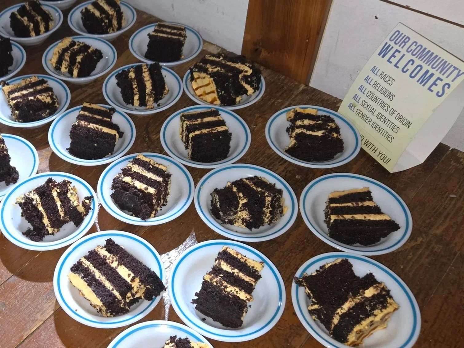 Several slices of Black Forest cake on small white plates with blue rim, displayed on a wooden table. A sign nearby reads 'Our Community Welcomes' with statements about inclusivity for all races, religions, nationalities, sexual and gender identities.