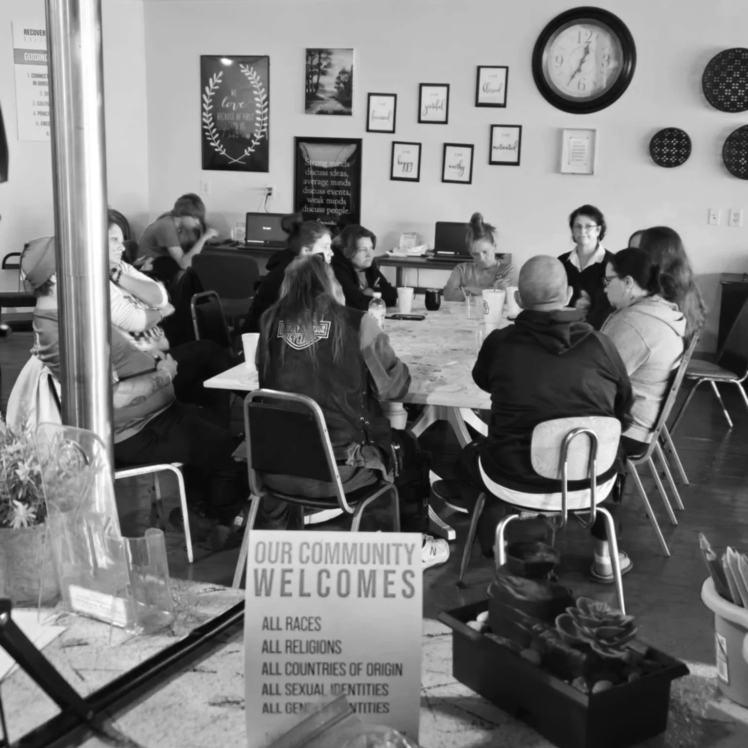 A group of people sitting around a long table in a community center or meeting room. There are framed pictures and a clock on the wall. A sign on the table reads, 'Our community welcomes all races, all religions, all countries of origin, all sexual identities, all genders.'