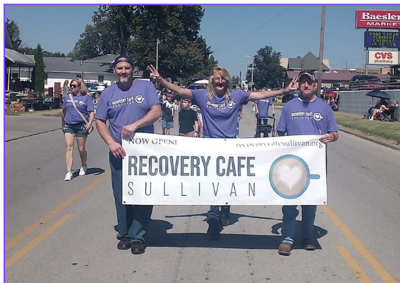 Three people in blue shirts are holding a banner that reads 'Recovery Cafe Sullivan' while standing in the middle of a street during a parade. Other parade participants and spectators are visible in the background.