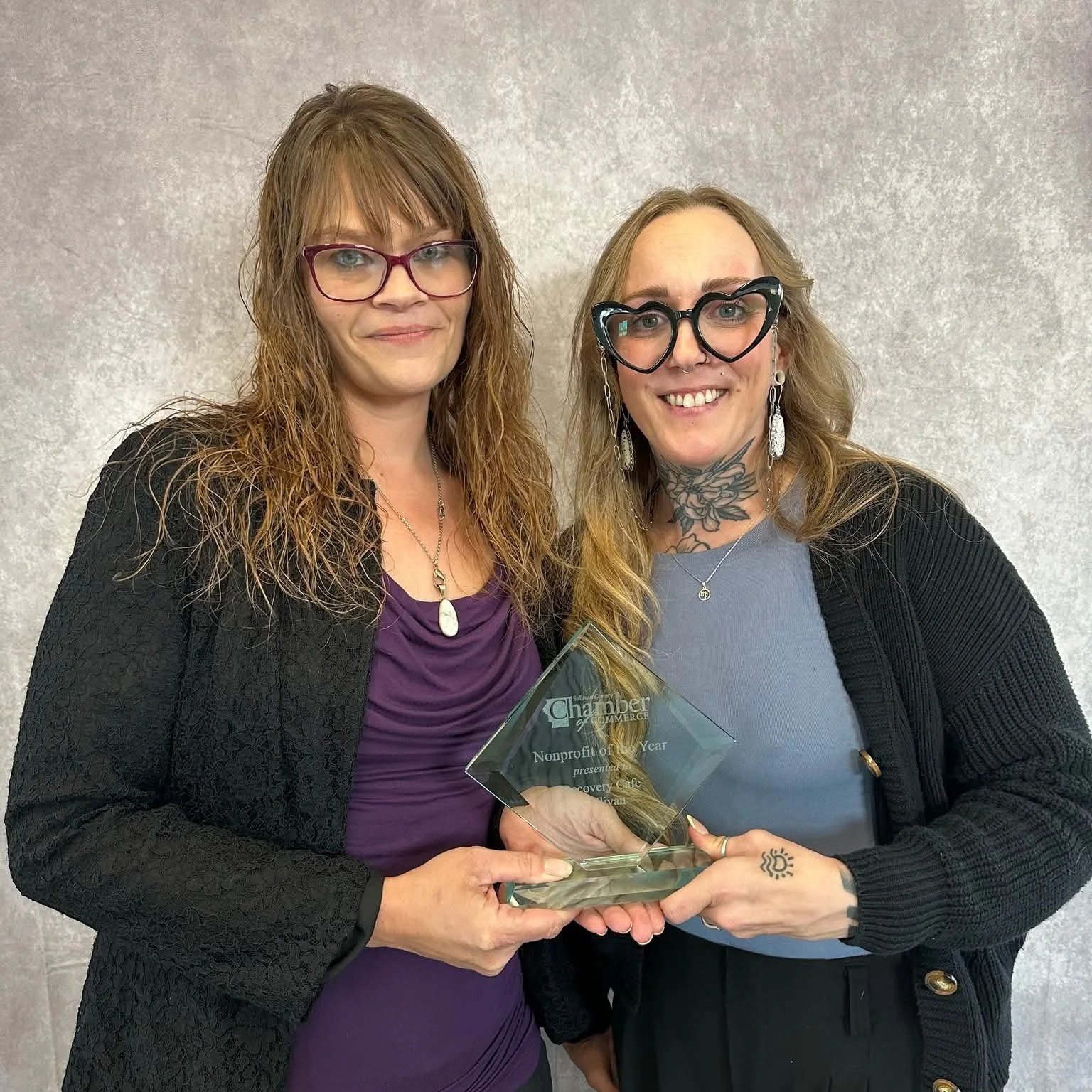 Two women holding an award trophy and smiling at the camera. One woman has red hair and purple glasses, the other has blond hair, heart-shaped glasses, and tattoos.
