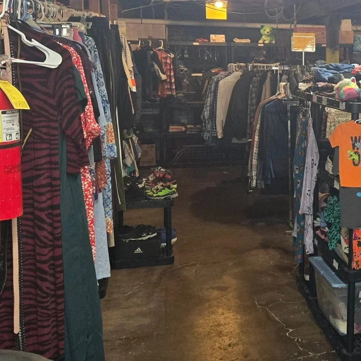 Clothing racks inside a thrift store, with various shirts, pants, and accessories on display, in a dimly lit space with a concrete floor.