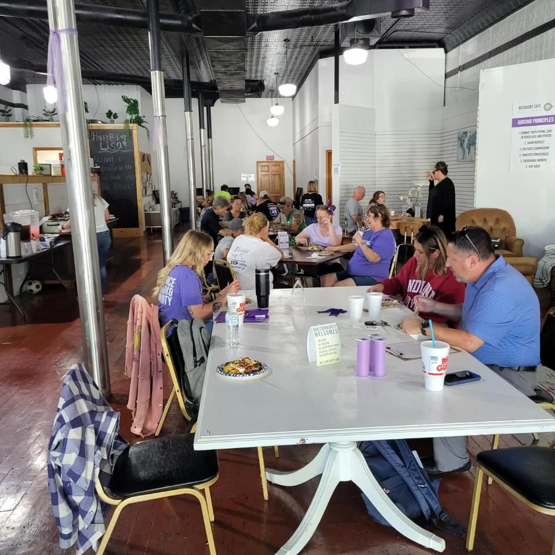 Interior of a cafe or community center with people sitting at tables, some eating and others using mobile devices, large white walls, a blackboard, and a sign on the wall, with a mix of casual seating and a cozy armchair.