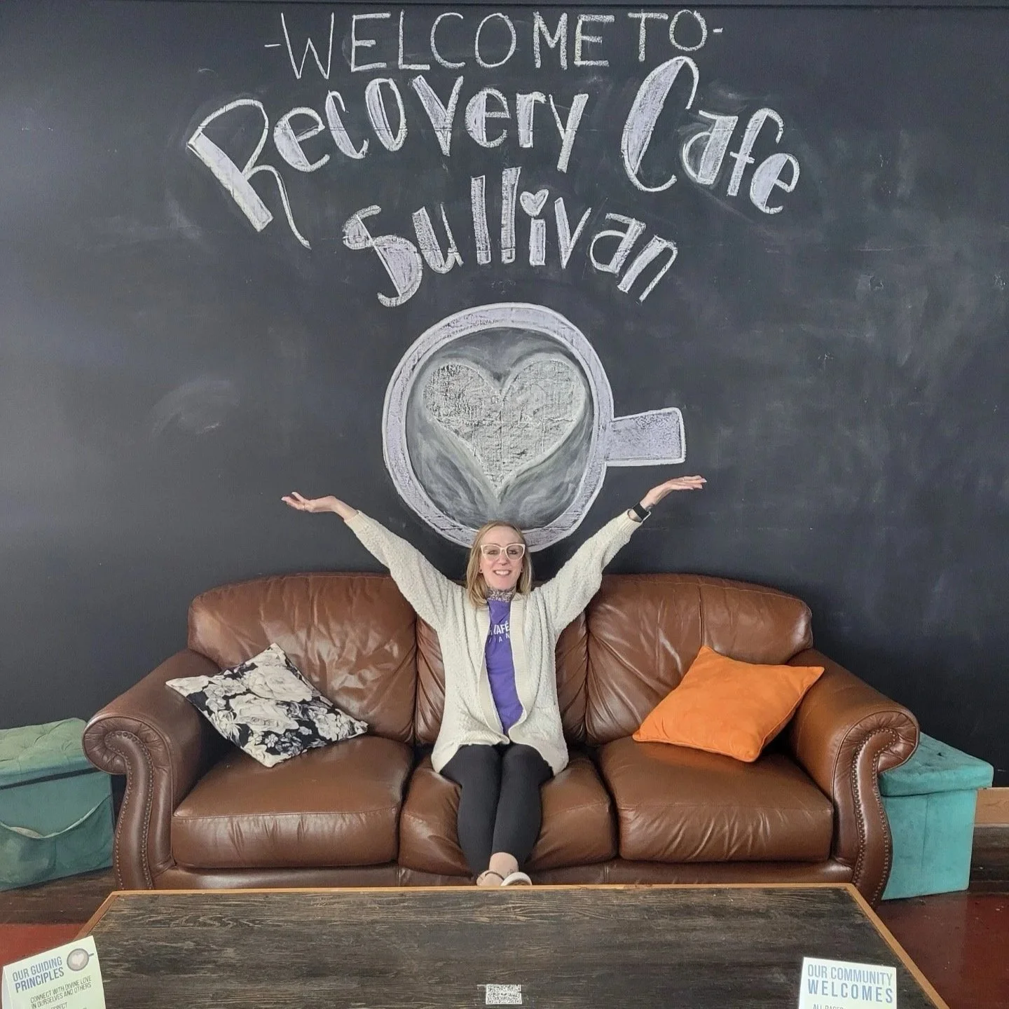 A woman sitting on a brown leather couch with her arms raised, in front of a black chalkboard wall with a chalk drawing of a coffee cup with a heart in it, and the words 'Welcome to Recovery Cafe Sullivan' and some small decor pillows on the couch.