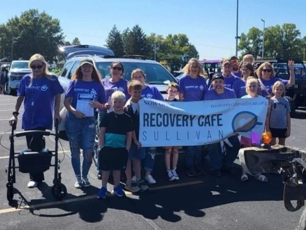 Group of people, including children and adults, holding a blue Recovery Cafe Sullivan banner in a parking lot on a sunny day.