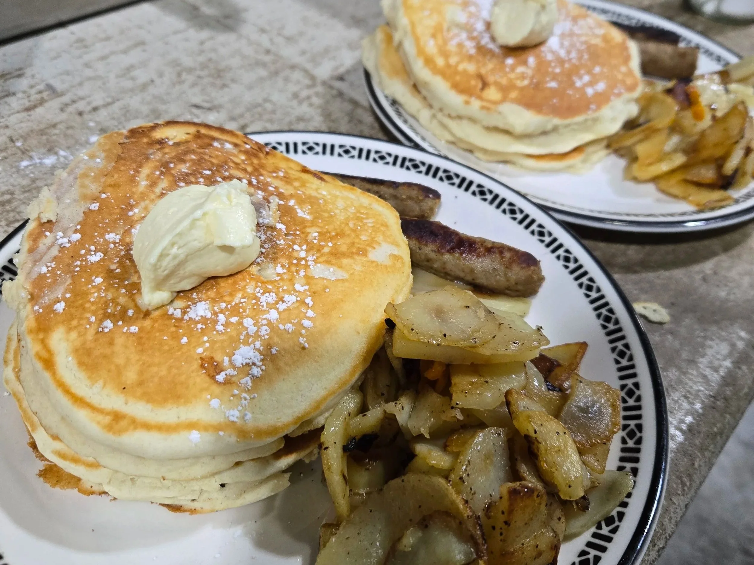 Pancakes topped with butter and powdered sugar served with sausage and hash browns on a patterned white plate