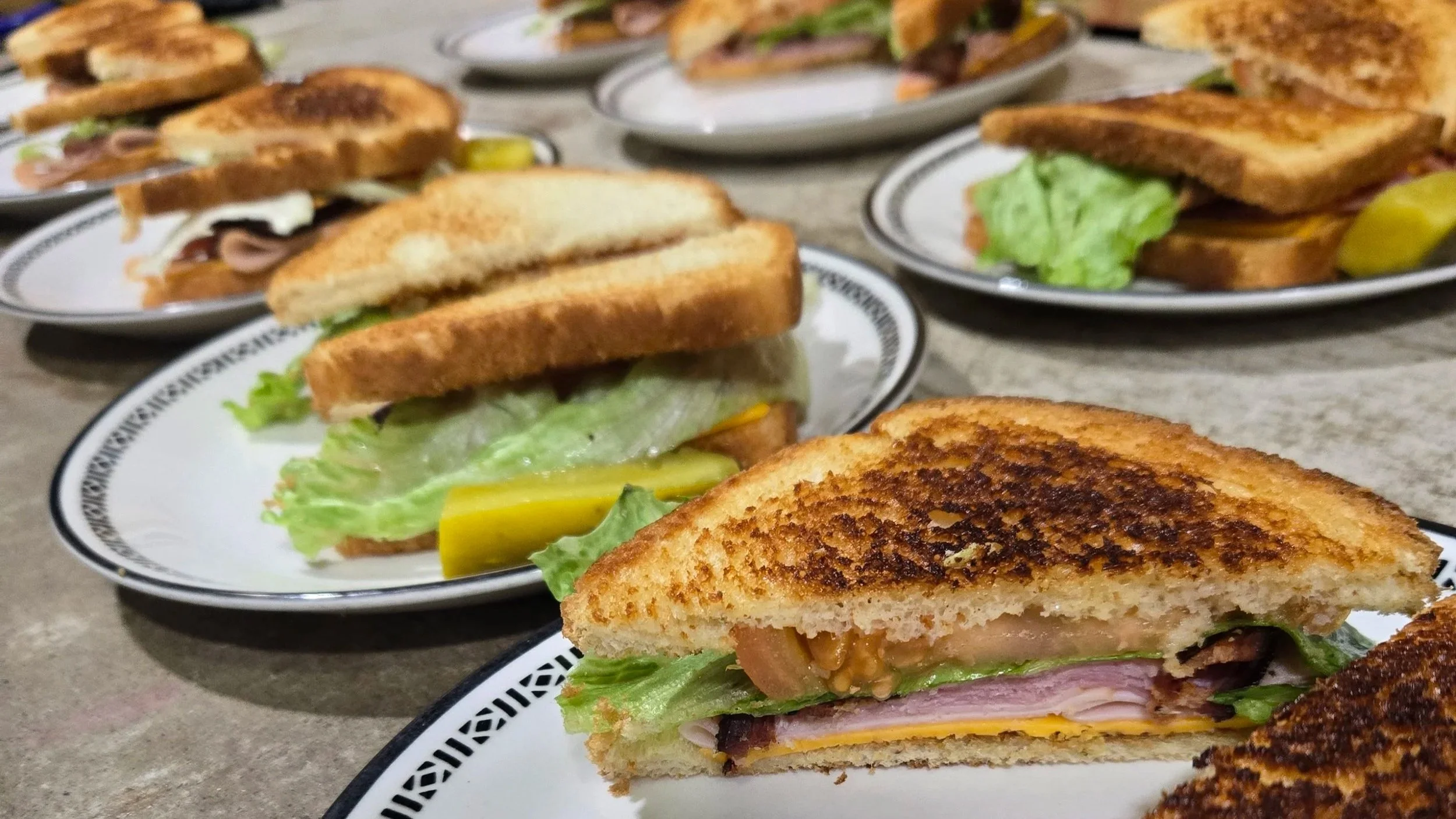 Close-up of grilled sandwiches with lettuce, tomato, cheese, and meat on white plates with black patterns, placed on a gray textured surface.