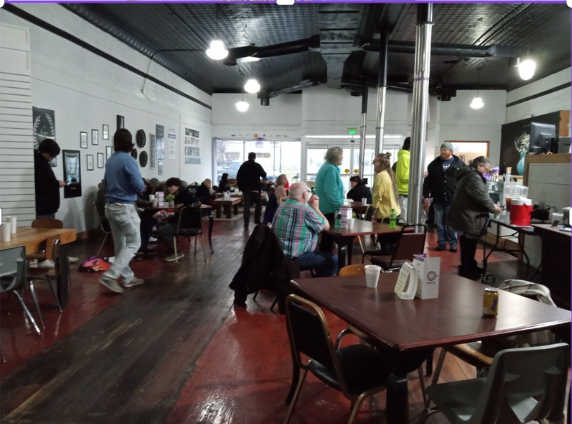 People inside a casual cafe or restaurant during daytime, some standing and others seated at tables, with a black ceiling, white walls, and wooden floors.