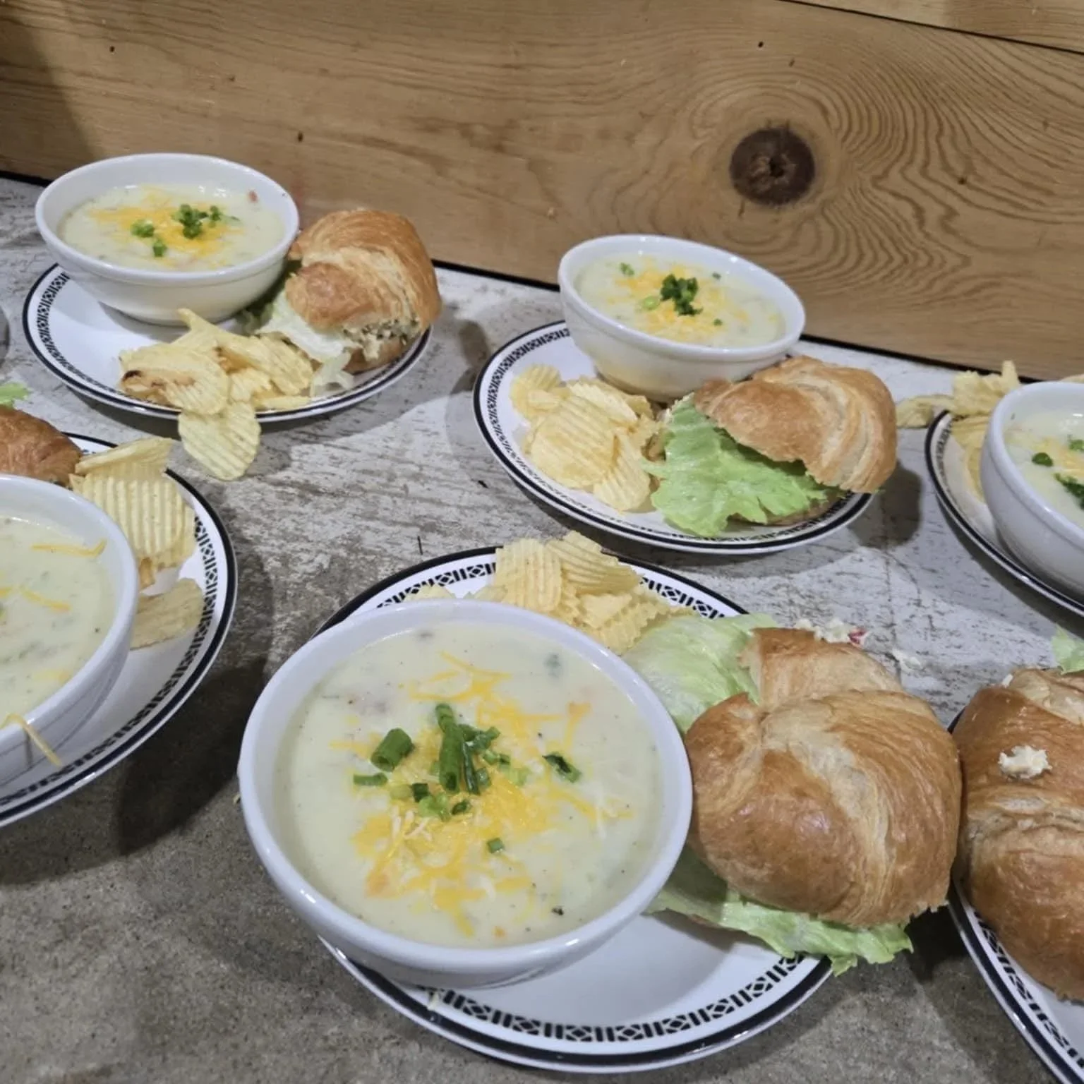 Several bowls of creamy soup garnished with cheese and green herbs, served with croissants, potato chips, and lettuce on plates.