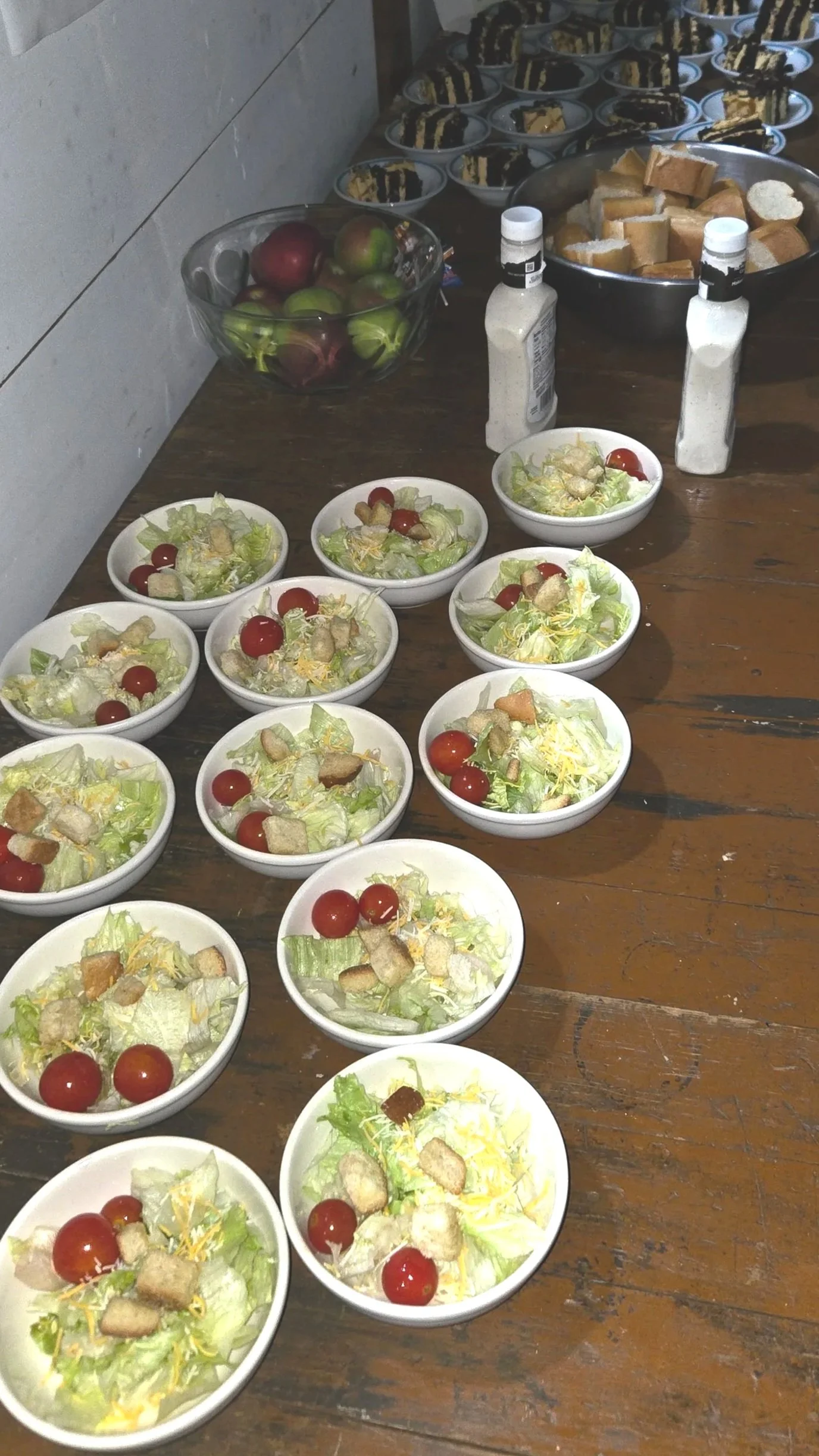 Multiple bowls of garden salad with lettuce, cherry tomatoes, shredded cheese, and croutons on a wooden table, with additional food items and condiments in the background.