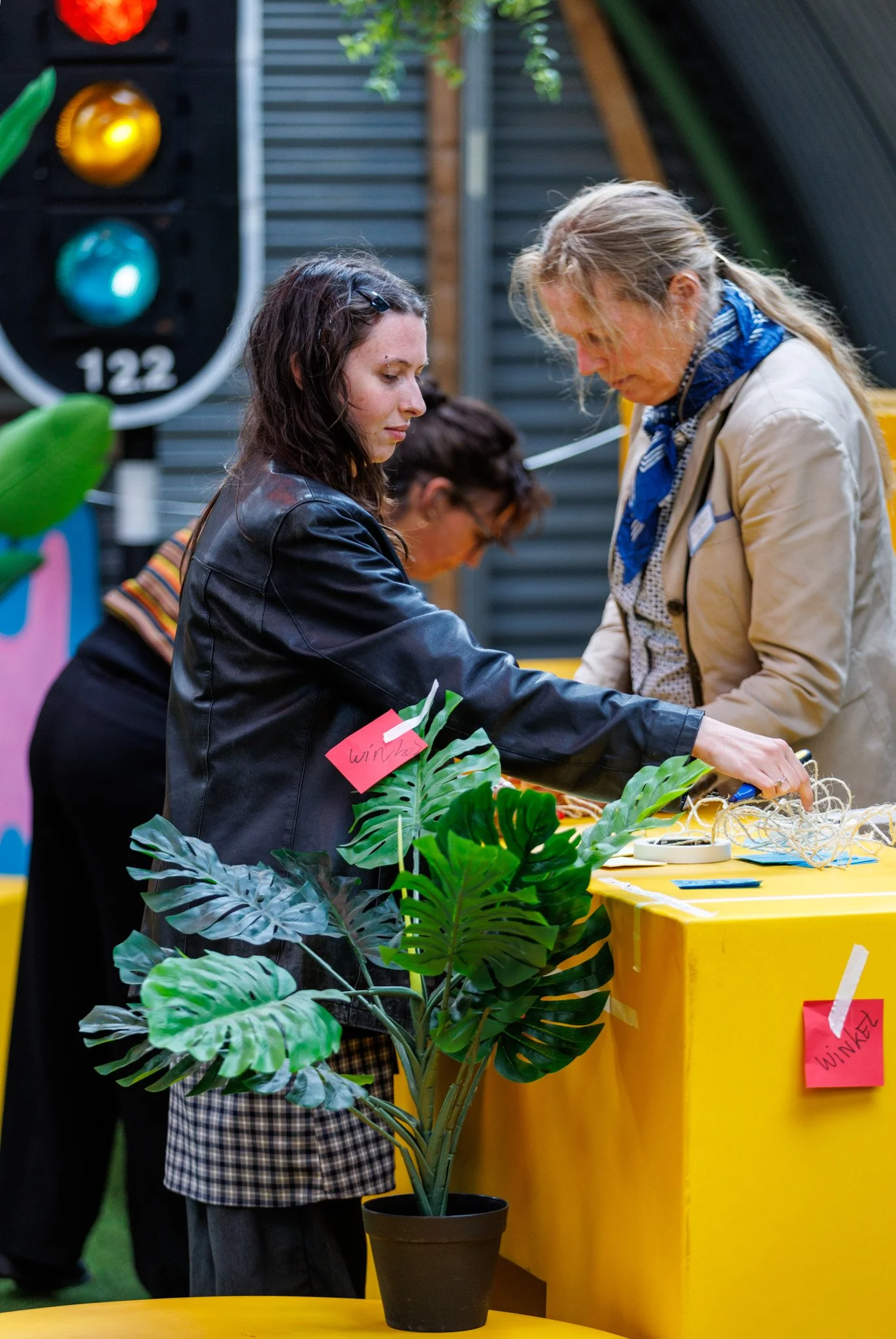 Three women gathered around a table with a potted monstera plant and flowers, at an indoor event with a yellow tablecloth and decorative signs.
