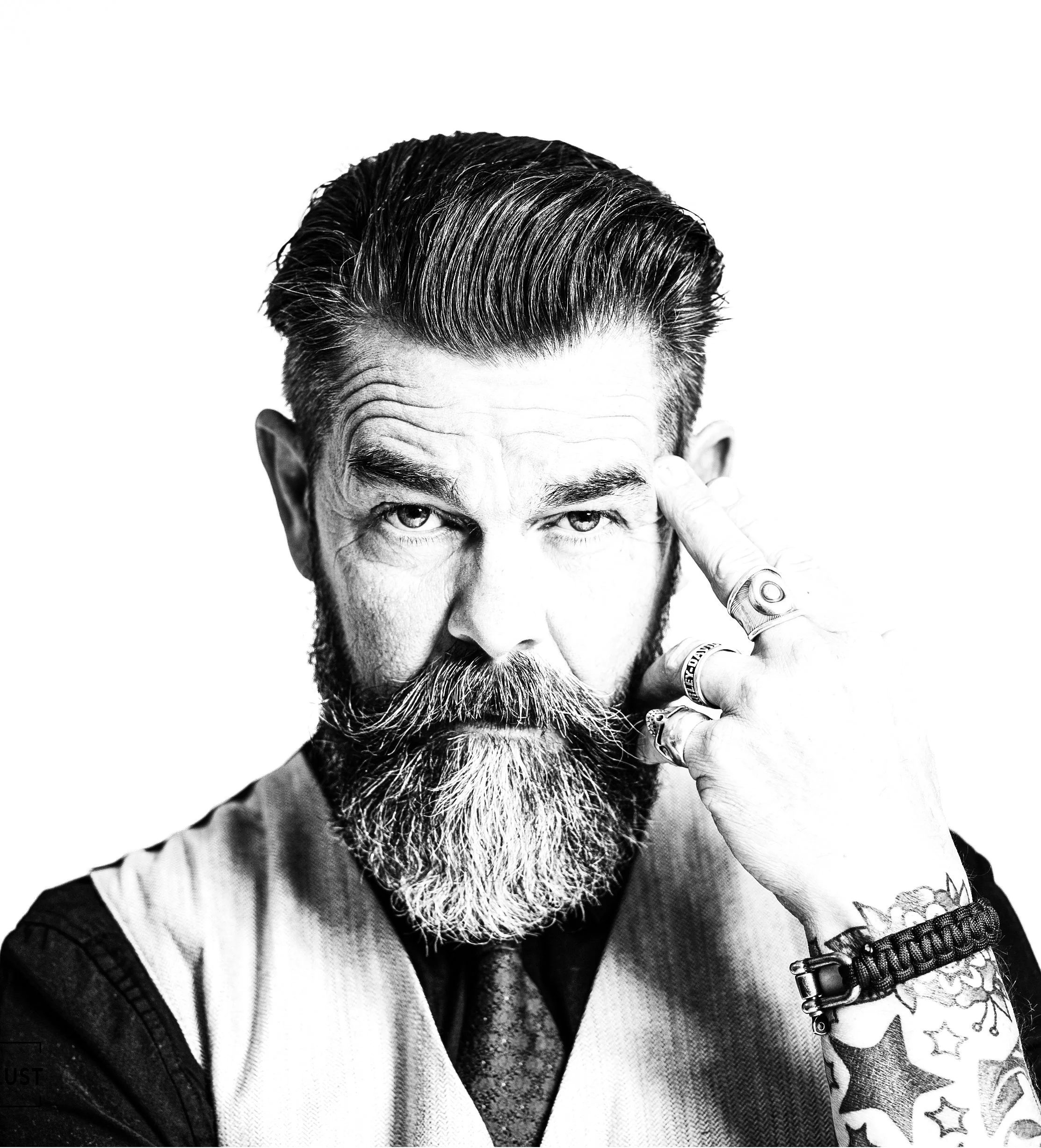 Black and white portrait of a bearded man with slicked-back hair, making a gesture near his temple with his right hand, which has tattoos and multiple rings, against a plain white background.
