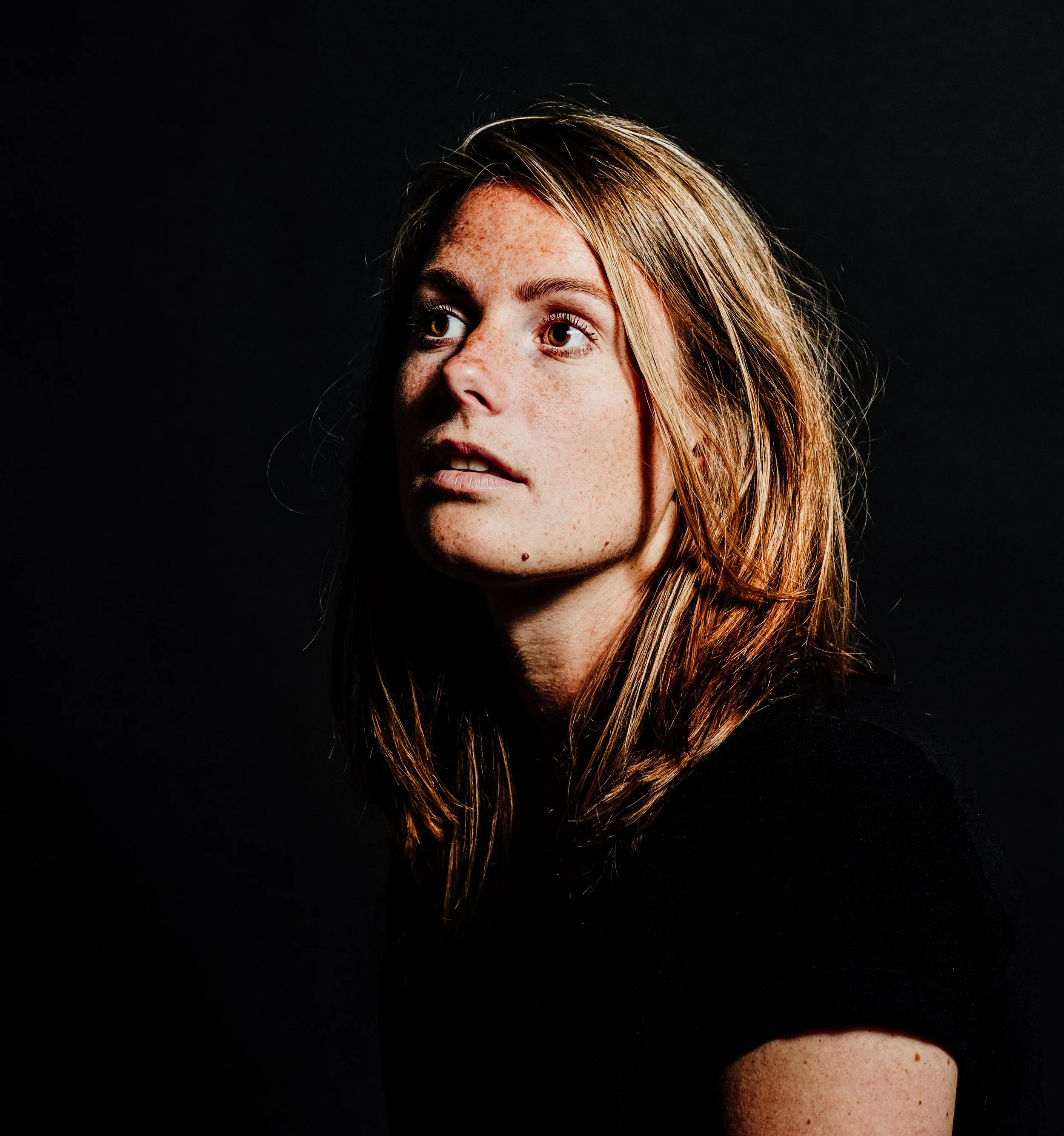 Portrait of a young woman with light brown hair and freckles against a black background.