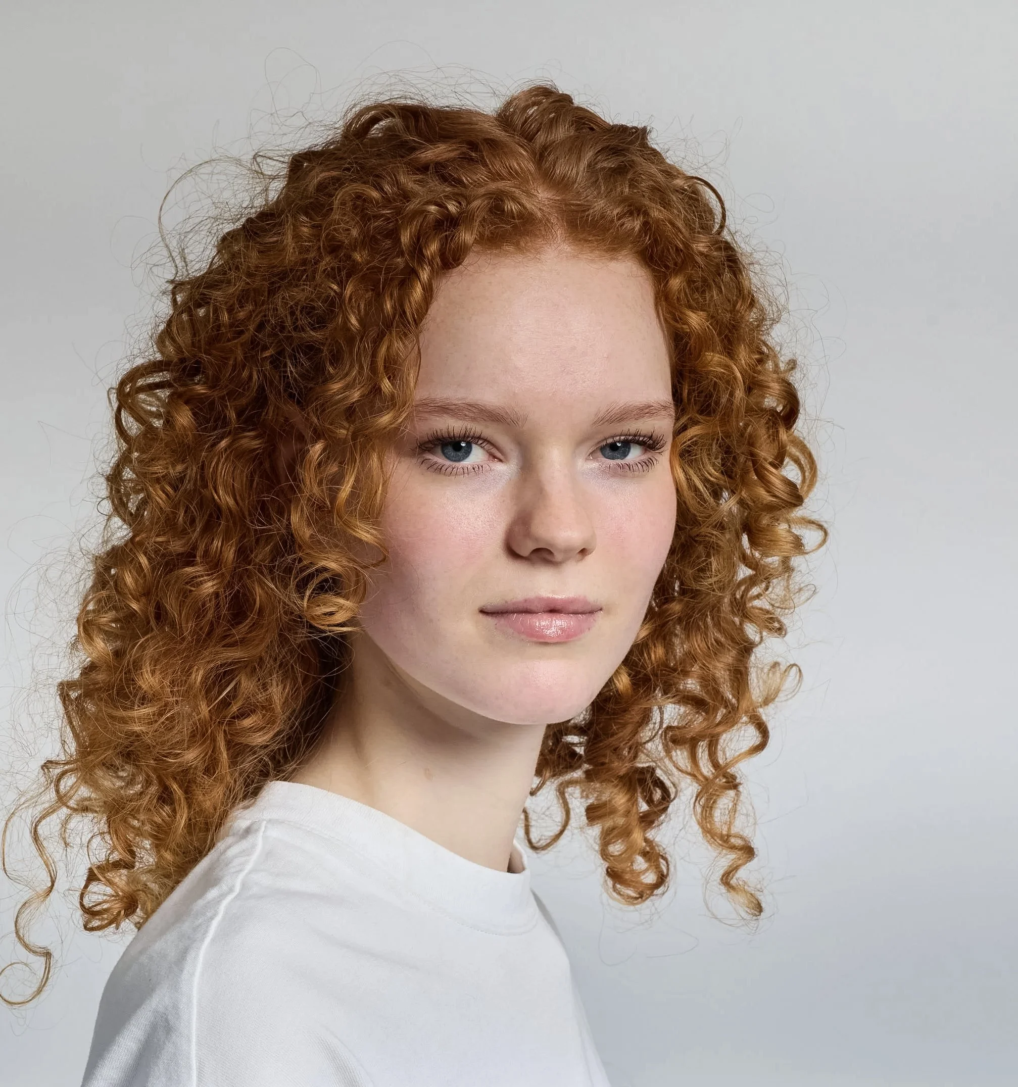 A young woman with curly red hair and blue eyes, wearing a white shirt, against a plain light gray background.