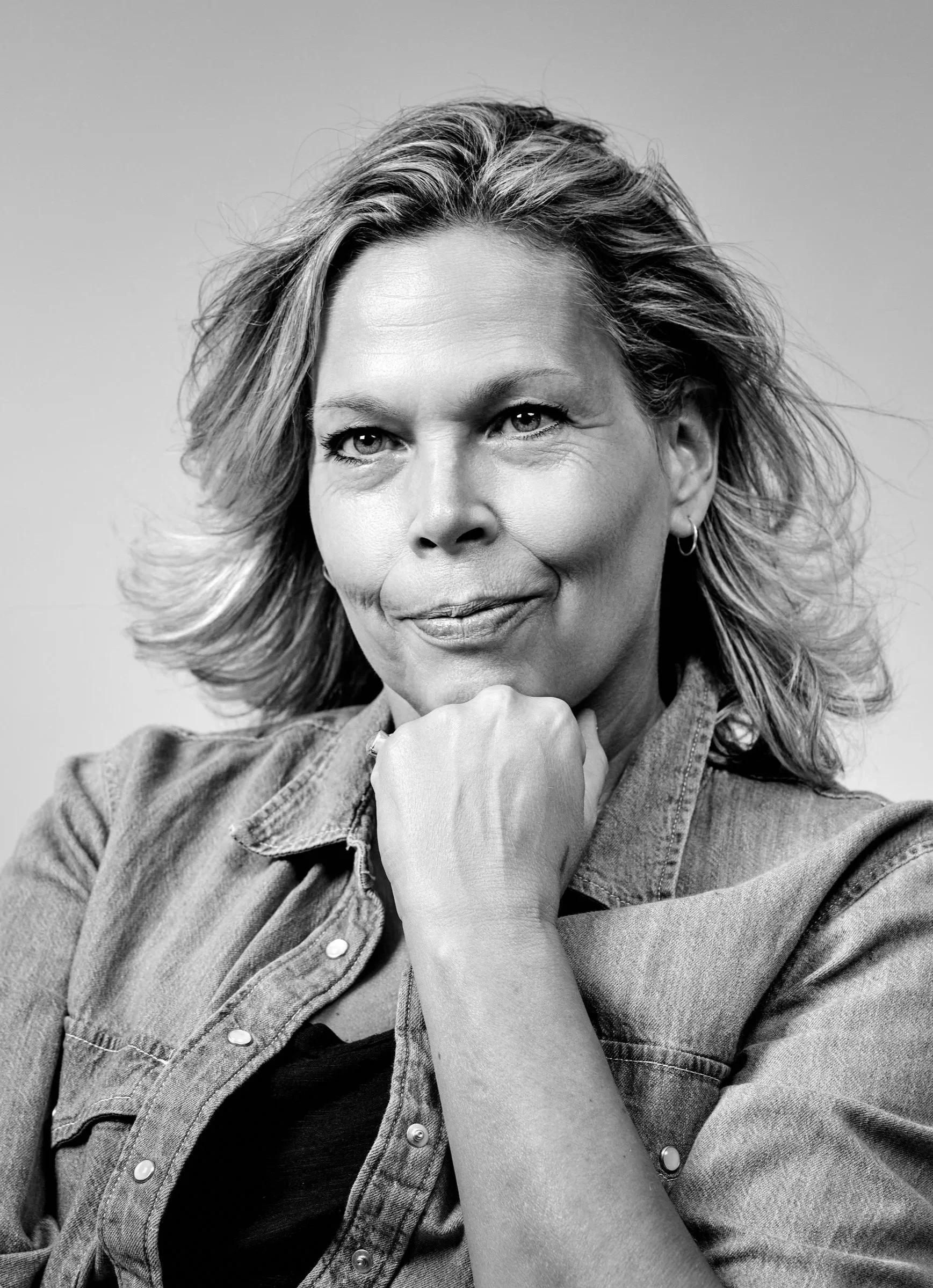 Zwarte en witte portretfoto van een vrouw met halflang haar die overwegend uit haar rechterkant kijkt, met haar hand onder haar chin, in een spijkerjack en zwarte top.
