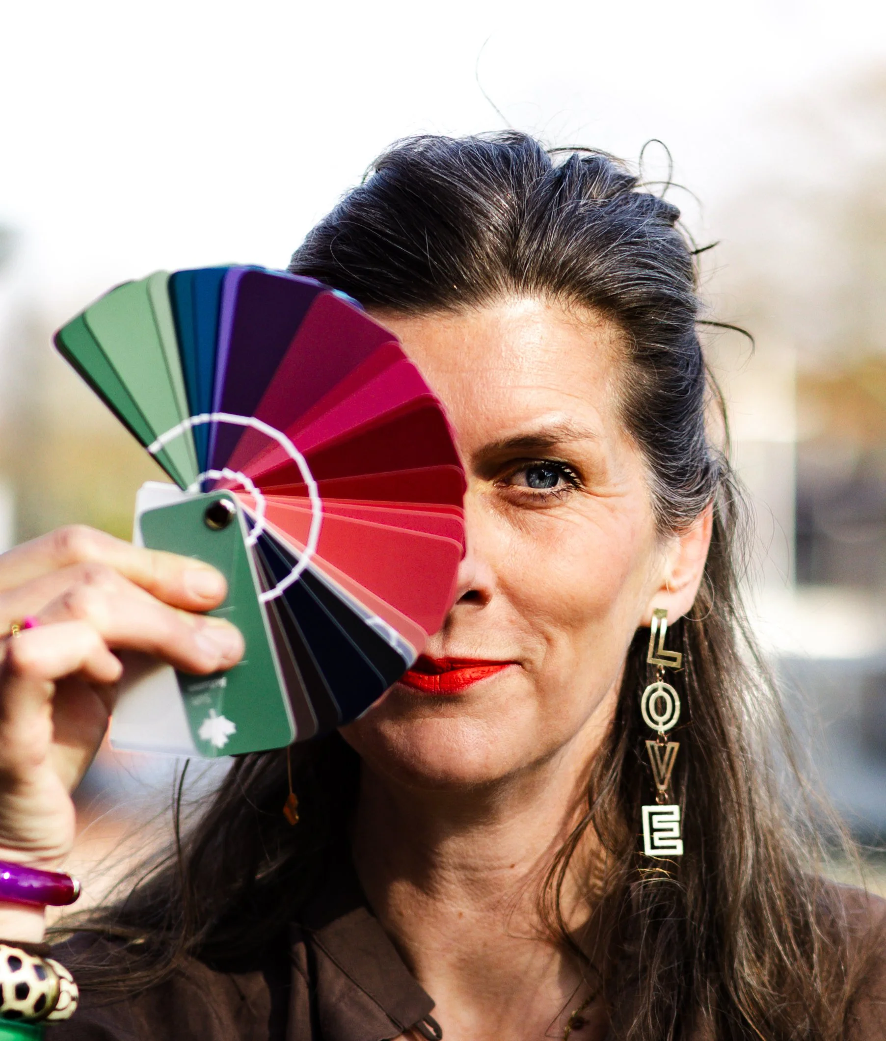 A woman with dark hair, blue eyes, and red lipstick holds a fan of color samples over her face, covering one eye.