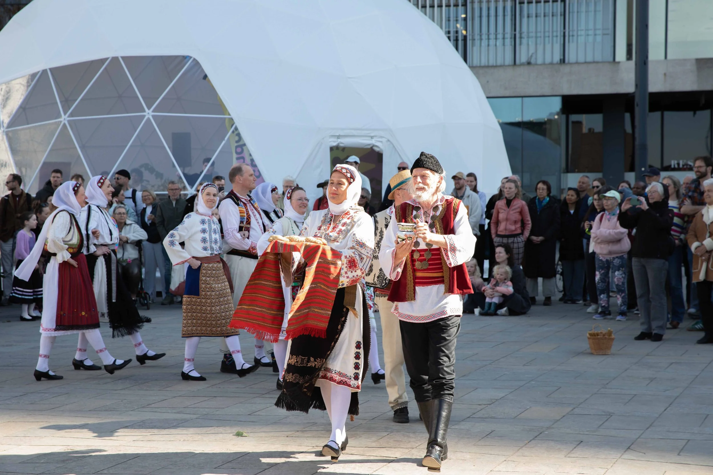 People dressed in traditional folk costumes performing and observing a cultural dance outdoors in front of a modern building with a geodesic dome structure.