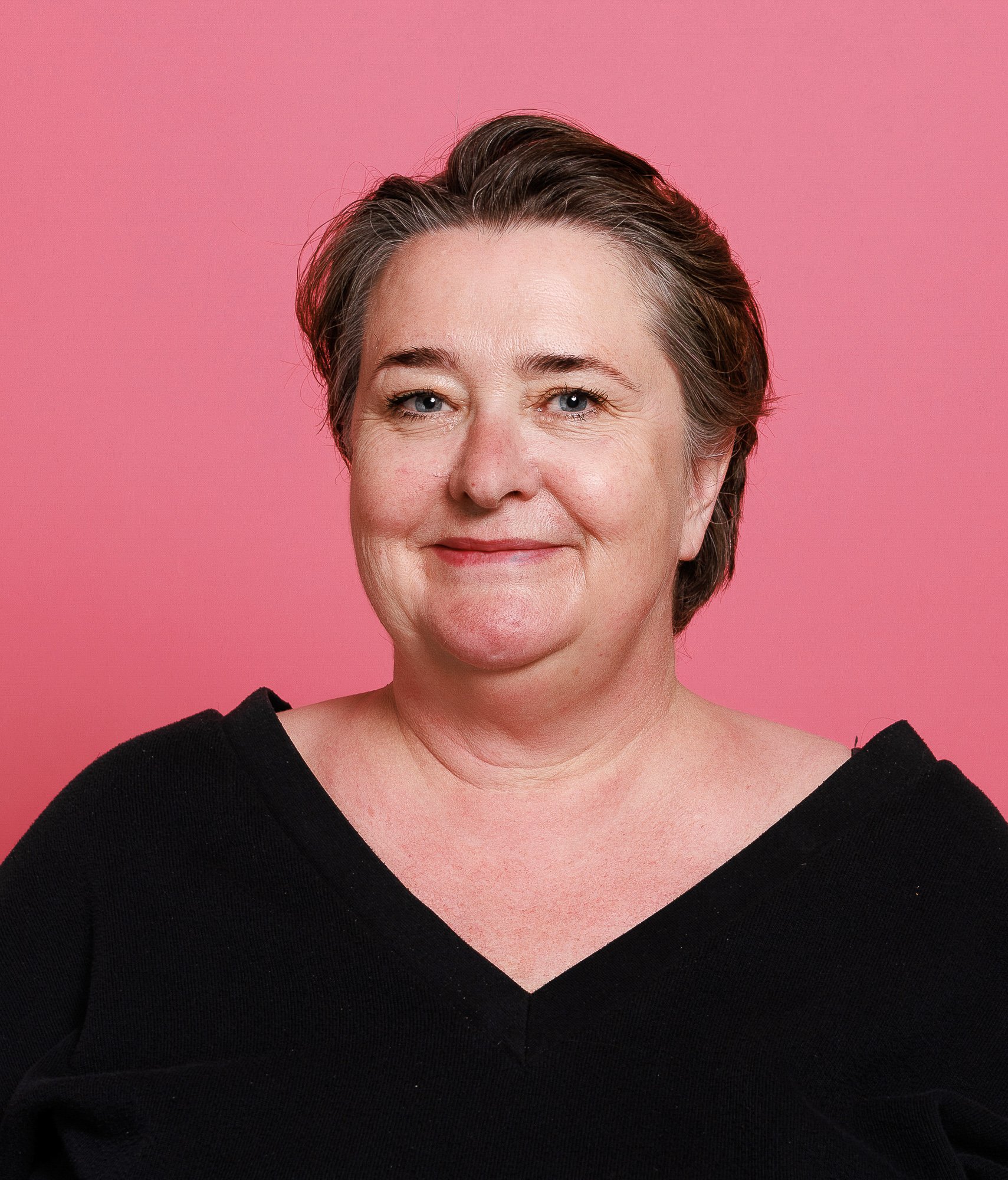 Portrait of a woman with short brown hair, wearing a black top, against a pink background.