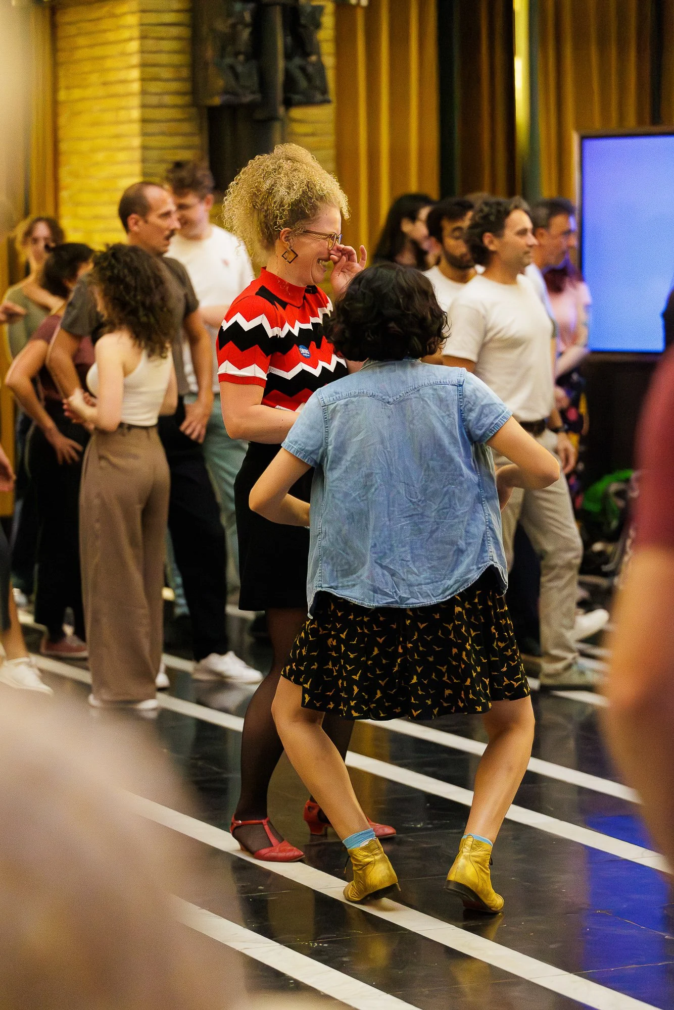 People dancing together in a lively indoor setting, wearing colorful clothing including a woman in a red, black, and white chevron patterned shirt and another woman in a denim and black skirt with bird patterns.