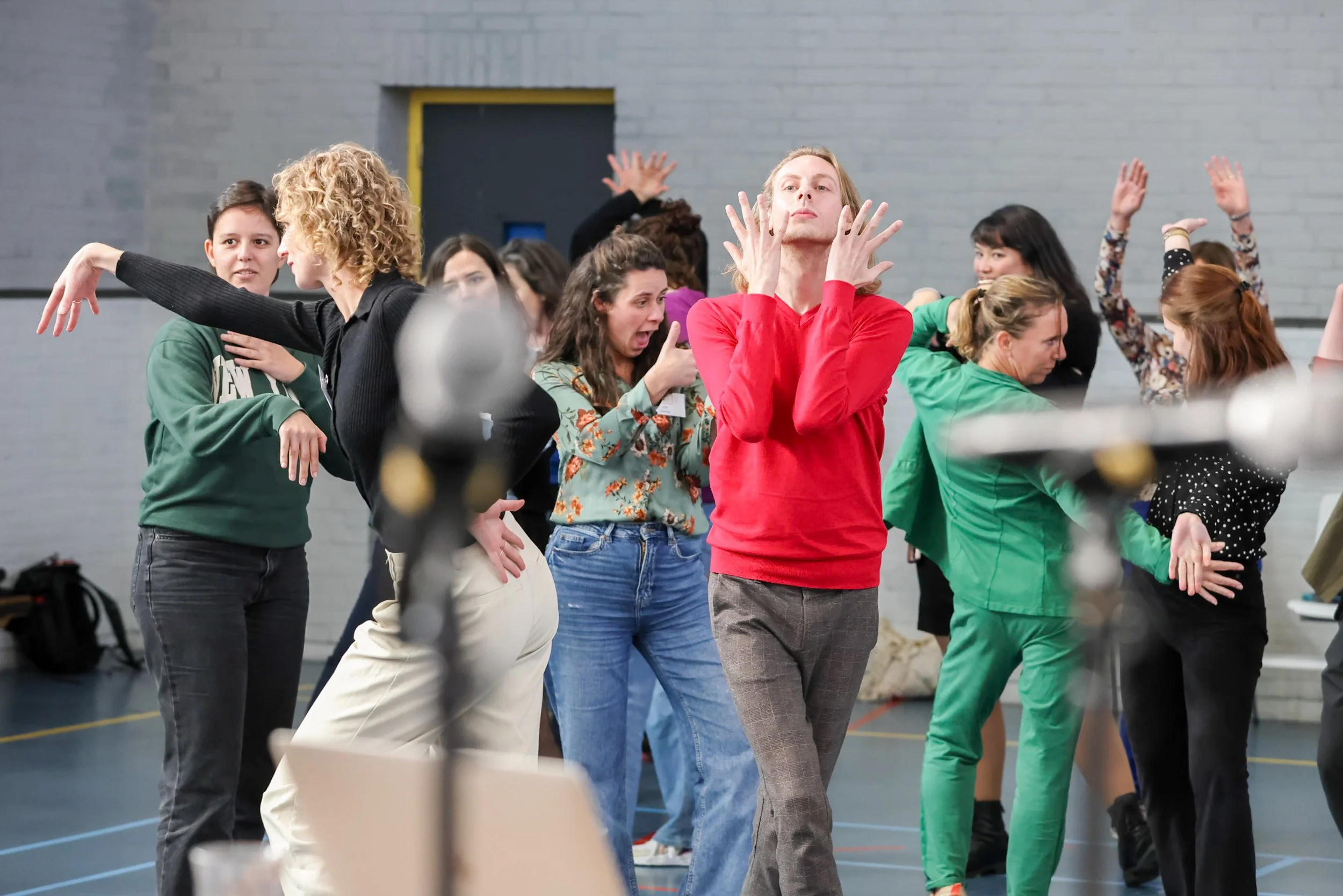 A group of people in a gym or dance studio practicing dance or theater moves, with some performers with outstretched arms and expressive poses.