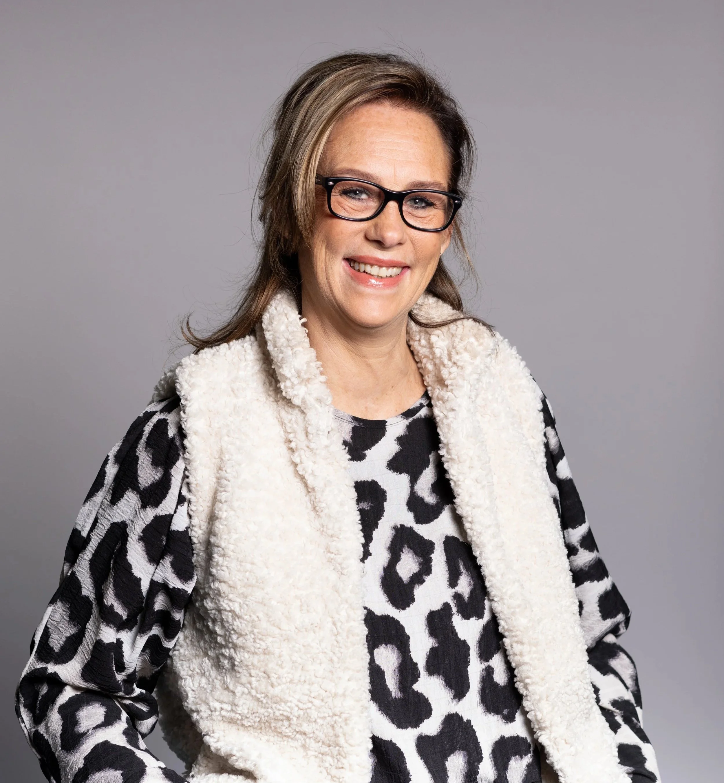 Happy woman wearing glasses and a black and white animal print outfit with a cream-colored fuzzy vest, standing against a gray background.