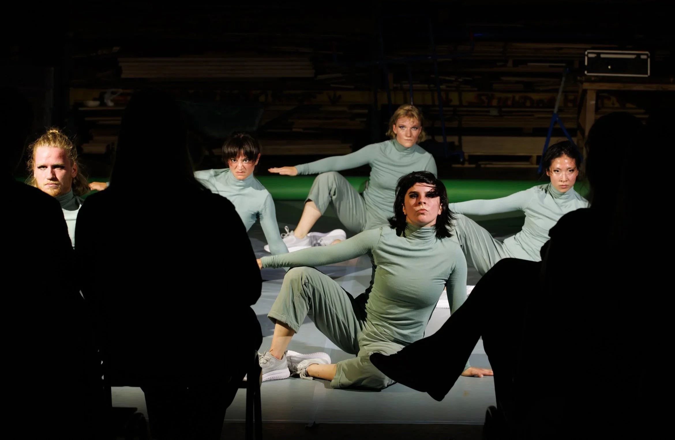Group of five performers in matching light green outfits on stage, with three audience members silhouetted in the foreground.
