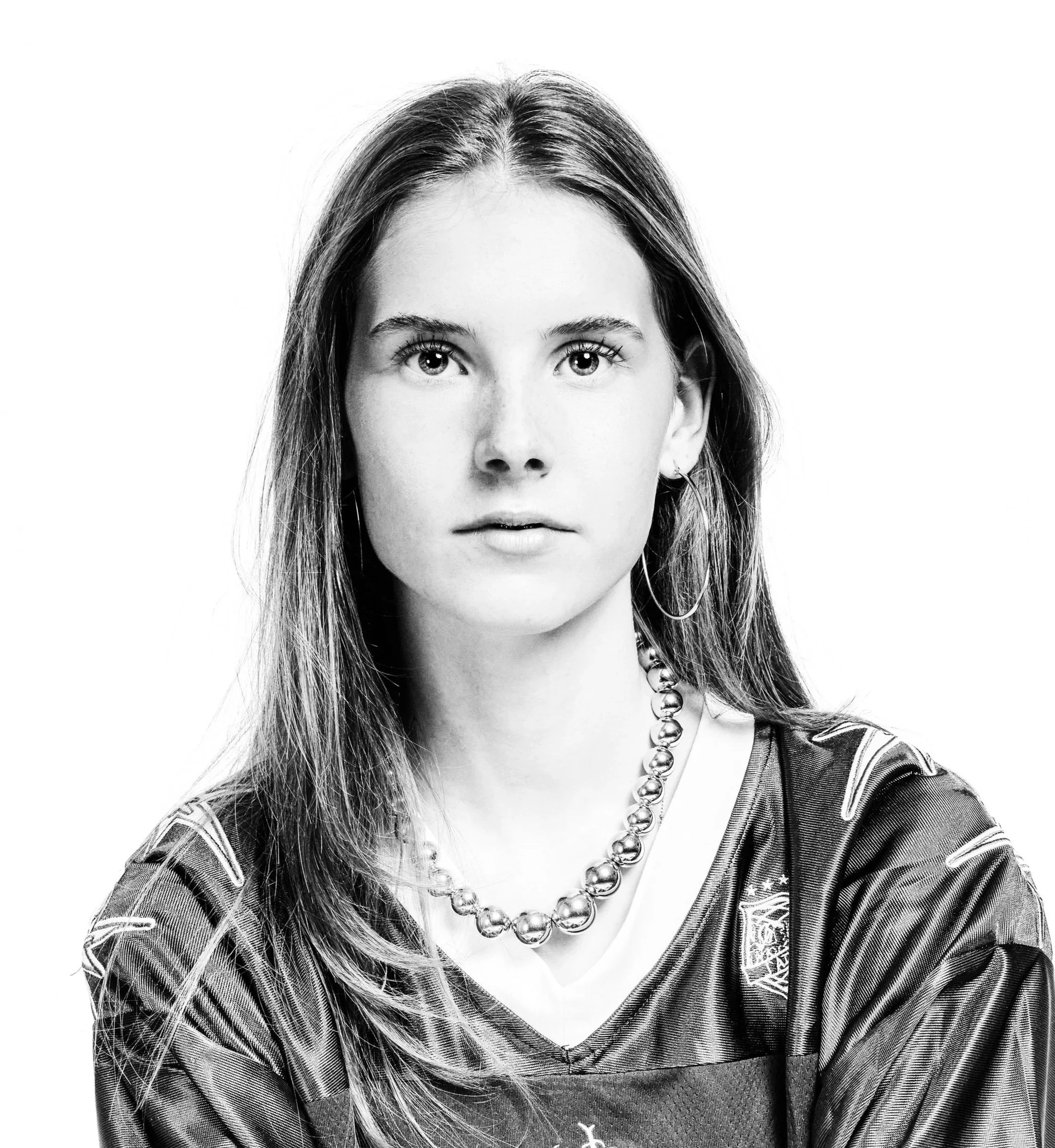 Black and white photo of a woman with straight hair, wearing a sports jersey, a pearl necklace, and hoop earrings, looking directly at the camera.