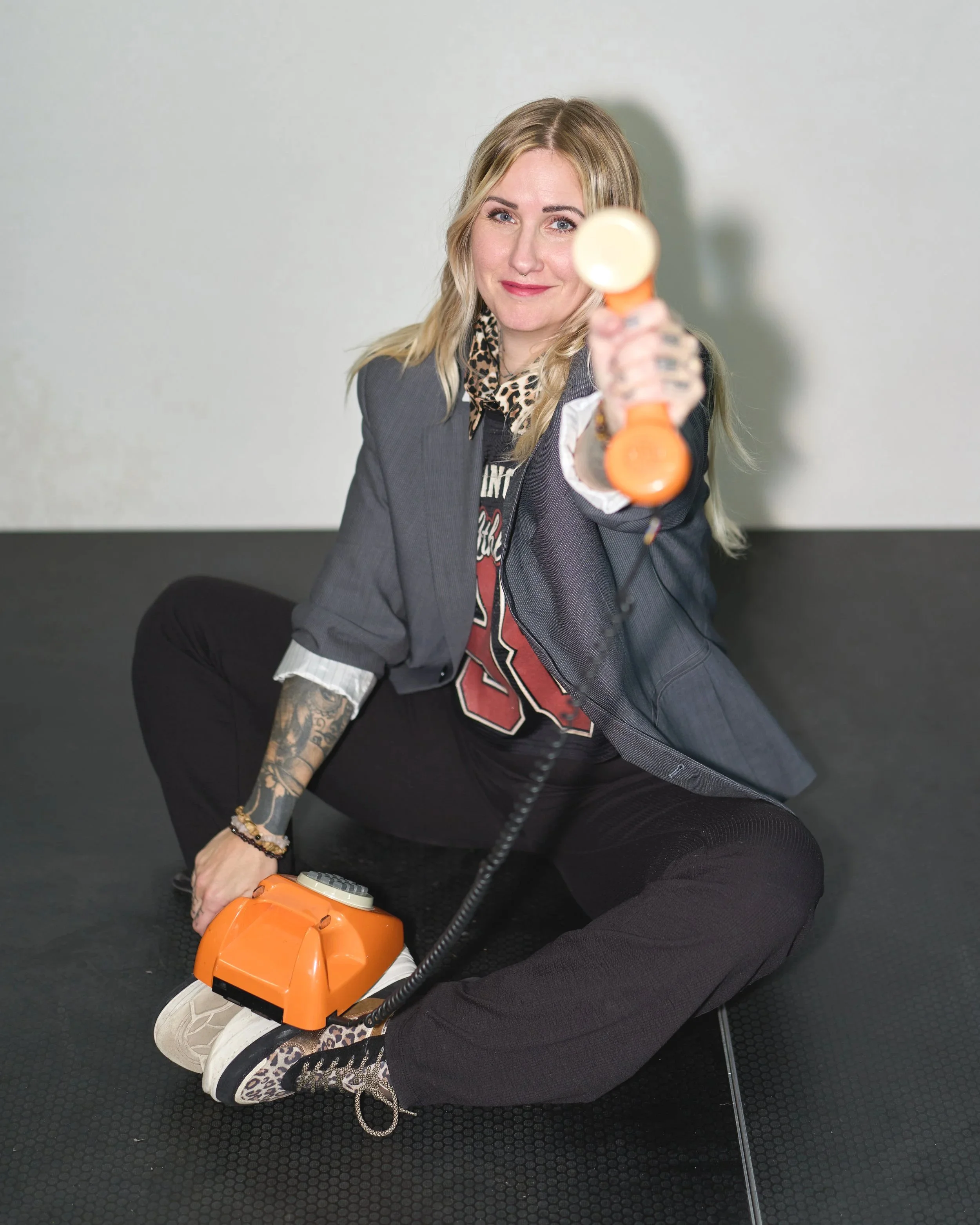 Woman with blonde hair and tattoos on her arm, wearing a blazer and leopard print shirt, holding an orange hairdryer pointing towards the camera, sitting on a black textured floor against a plain light-colored wall.