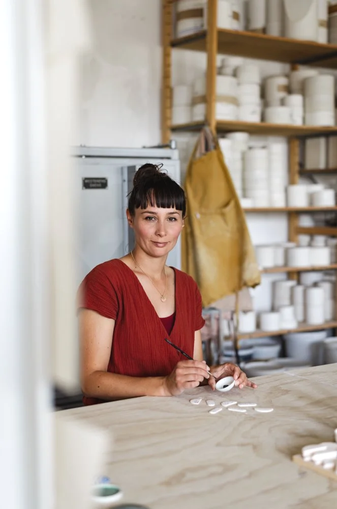 Woman with dark hair in a bun, wearing a red blouse, painting ceramic objects at a workshop with shelves of white ceramic pots in the background.