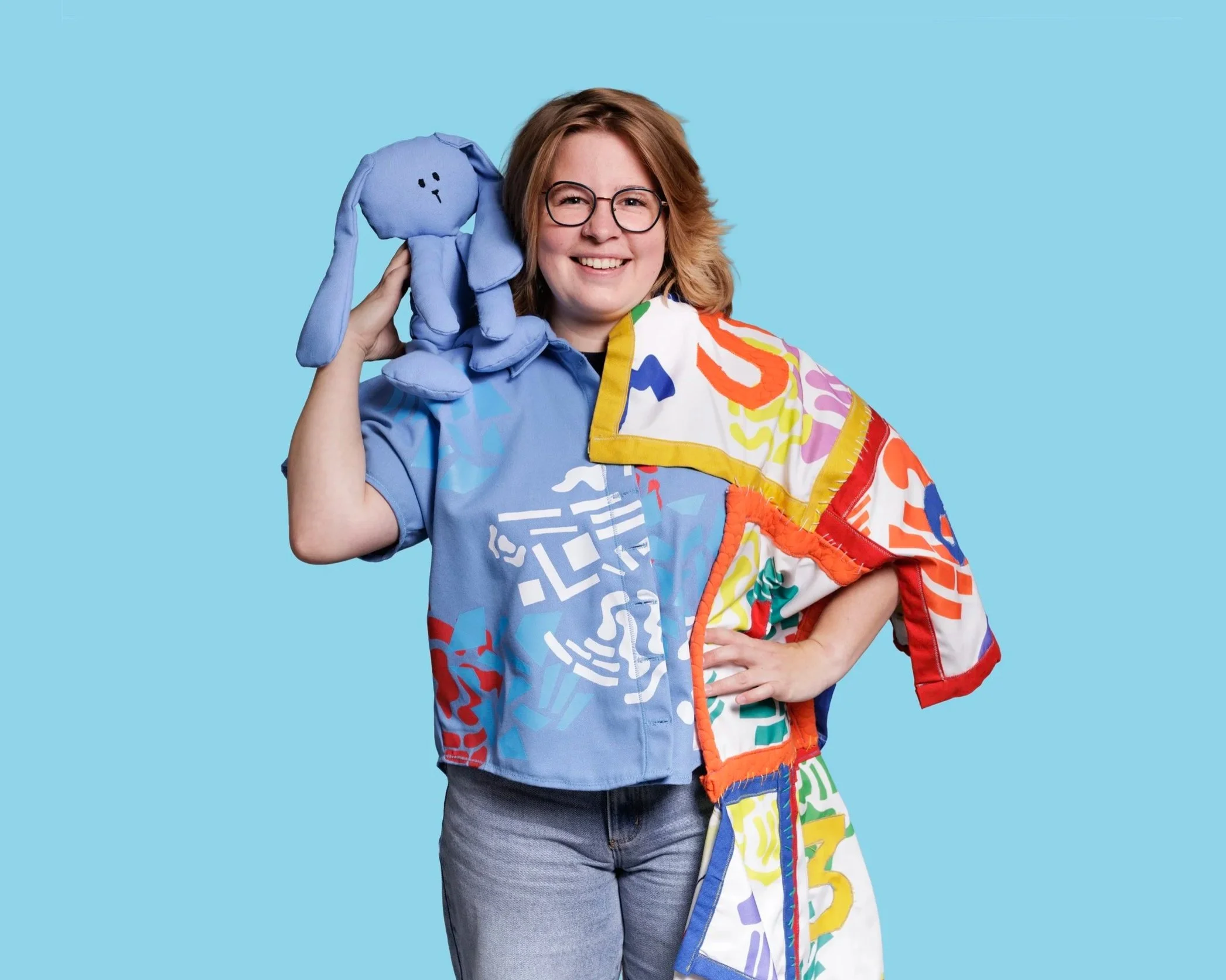 A woman with glasses, wearing a colorful patterned jacket and jeans, smiling while holding a blue stuffed animal to her ear, against a light blue background.