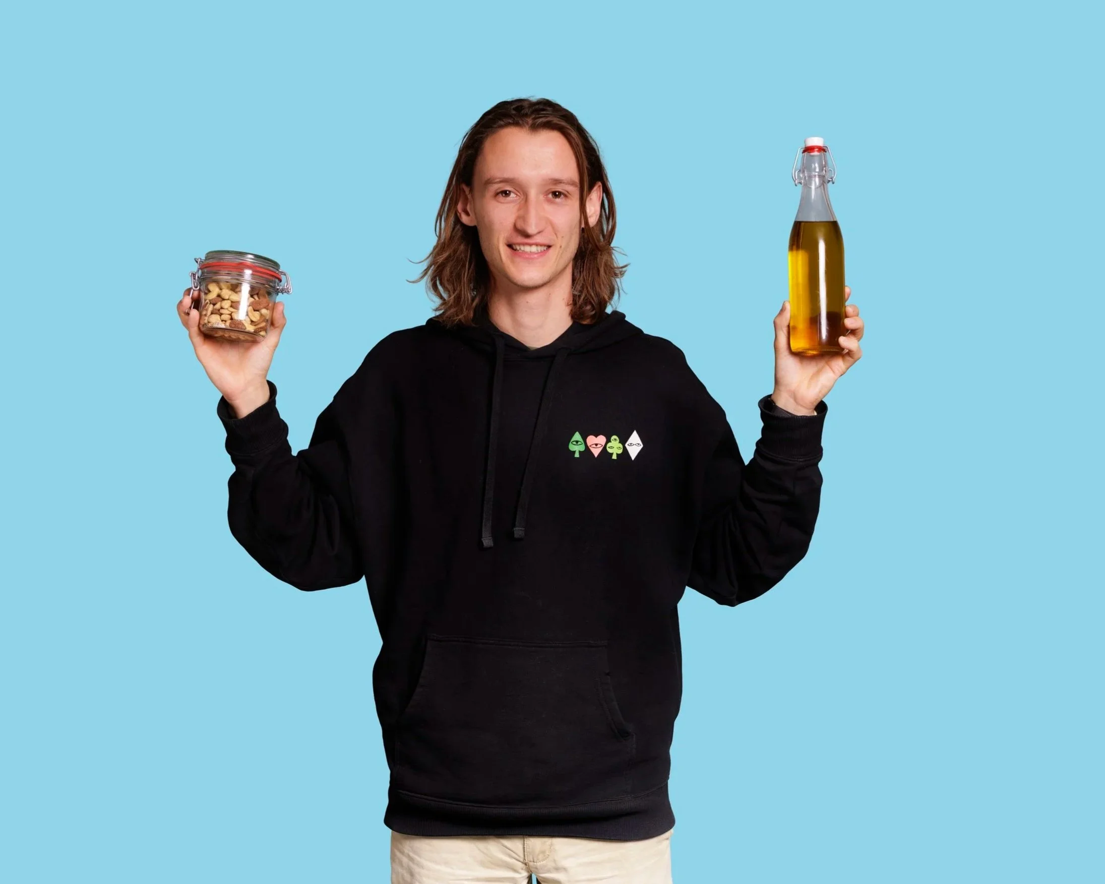 A young man with long brown hair in a black hoodie holding a jar of mixed nuts in one hand and a glass bottle of olive oil with a swing top in the other, standing against a light blue background.