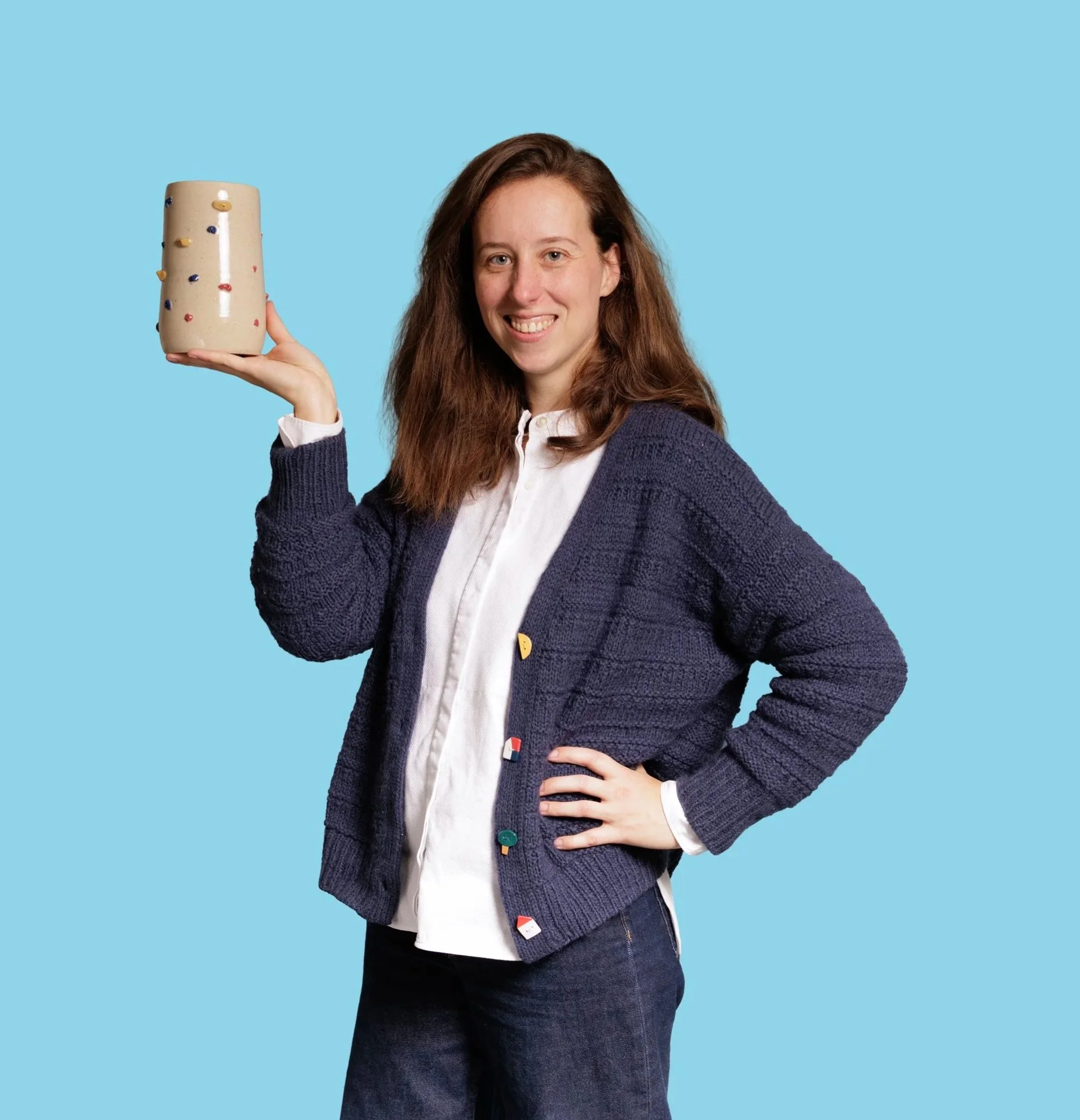 A woman with long brown hair smiling and holding a beige ceramic mug with colorful beads on a light blue background.
