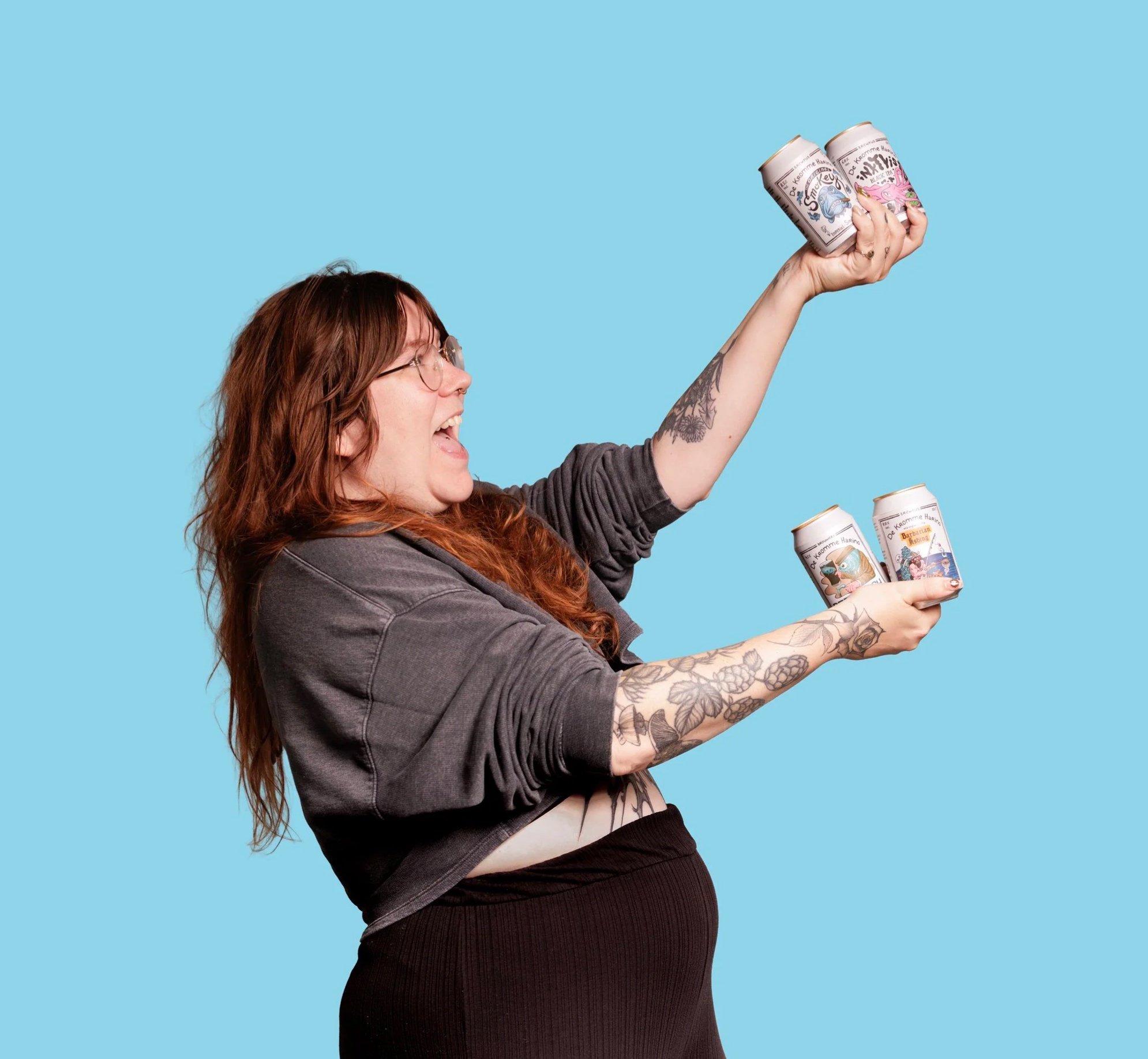 A woman with red hair and tattoos on her arm holding up cans of drink against a blue background.