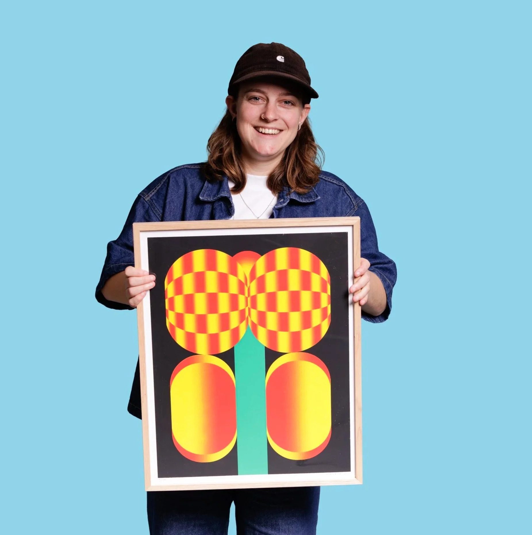 A person smiling, wearing a black cap and denim jacket, holding a colorful abstract artwork on a board, against a solid light blue background.