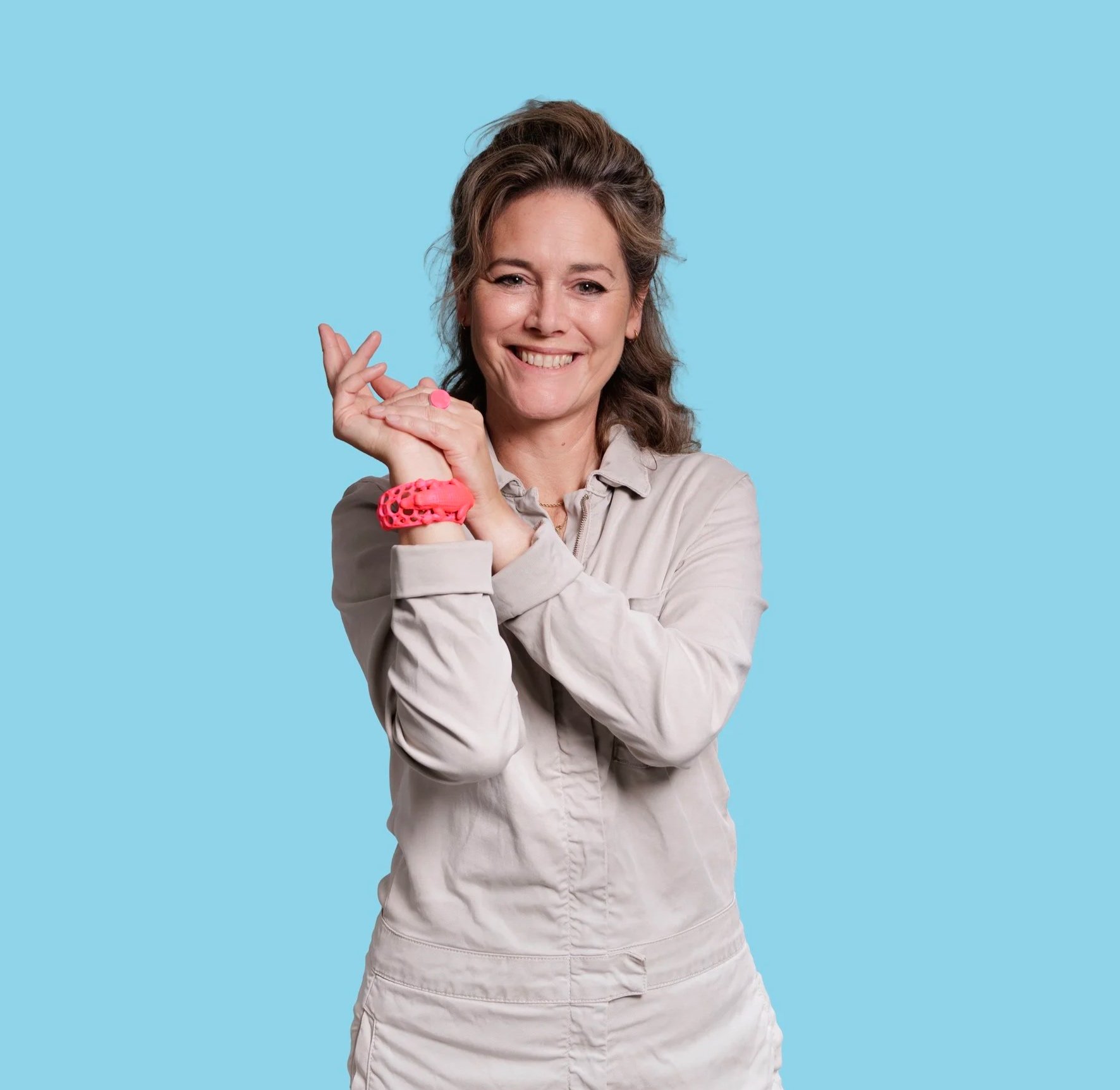 A woman with light brown, wavy hair, smiling, wearing a beige shirt, holding a pink item in her right hand, and standing against a solid light blue background.