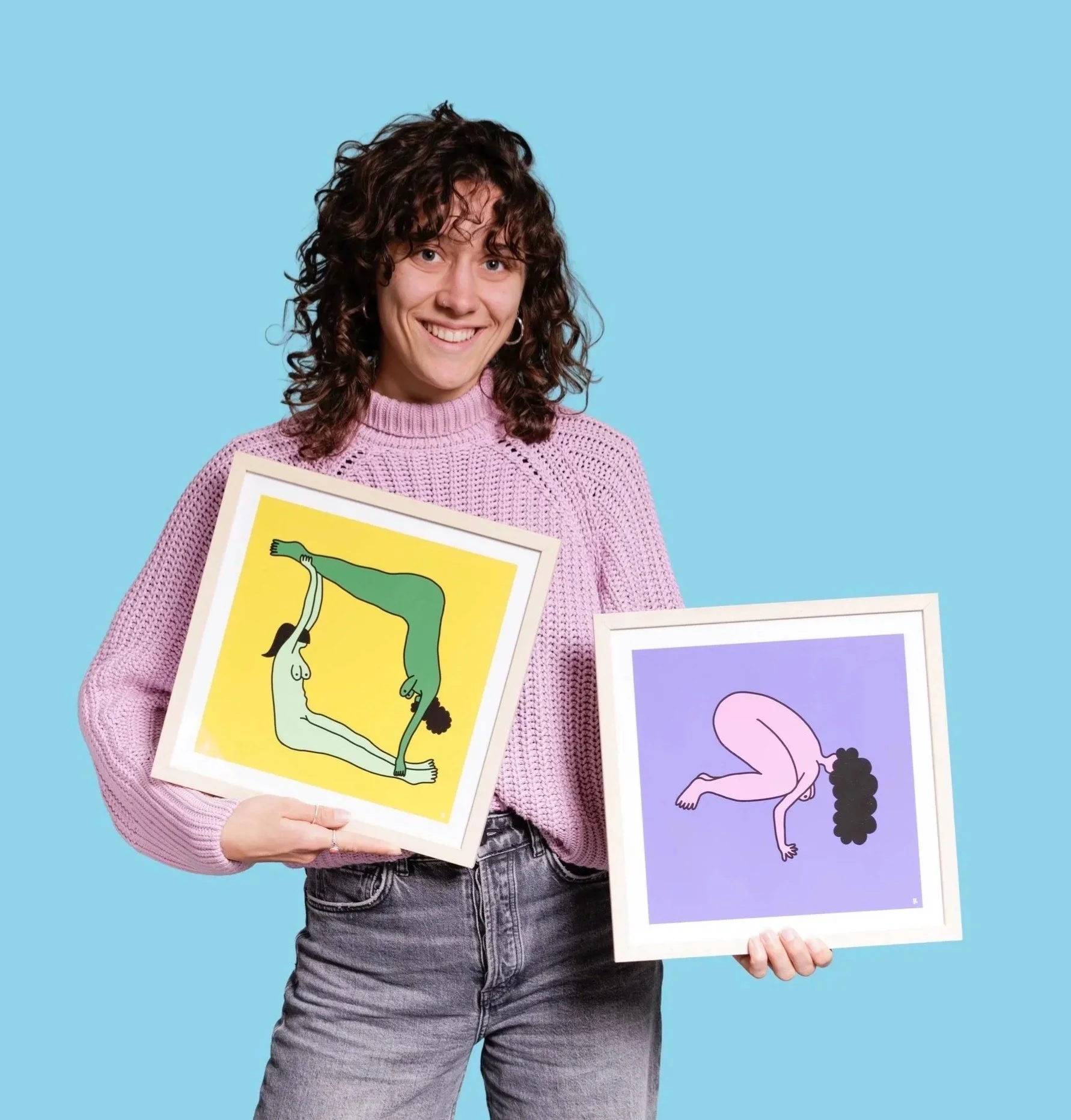 A smiling woman with curly brown hair holding two framed art prints with abstract, colorful illustrations against a solid light blue background.
