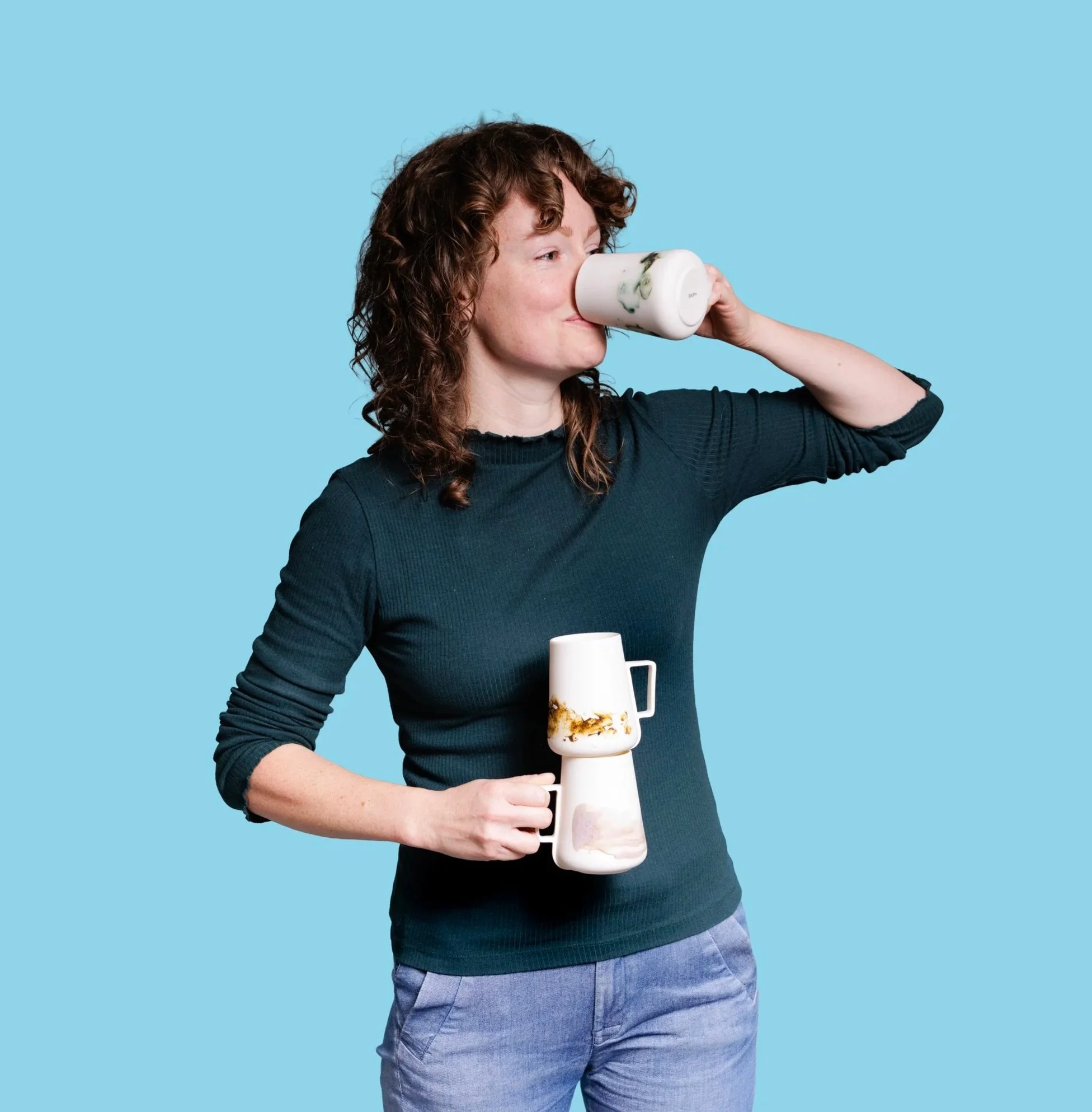 A woman with curly brown hair wearing a dark long-sleeve shirt holds a white mug with a floral design and a frappe glass layered with whipped cream and caramel sauce, standing against a light blue background.