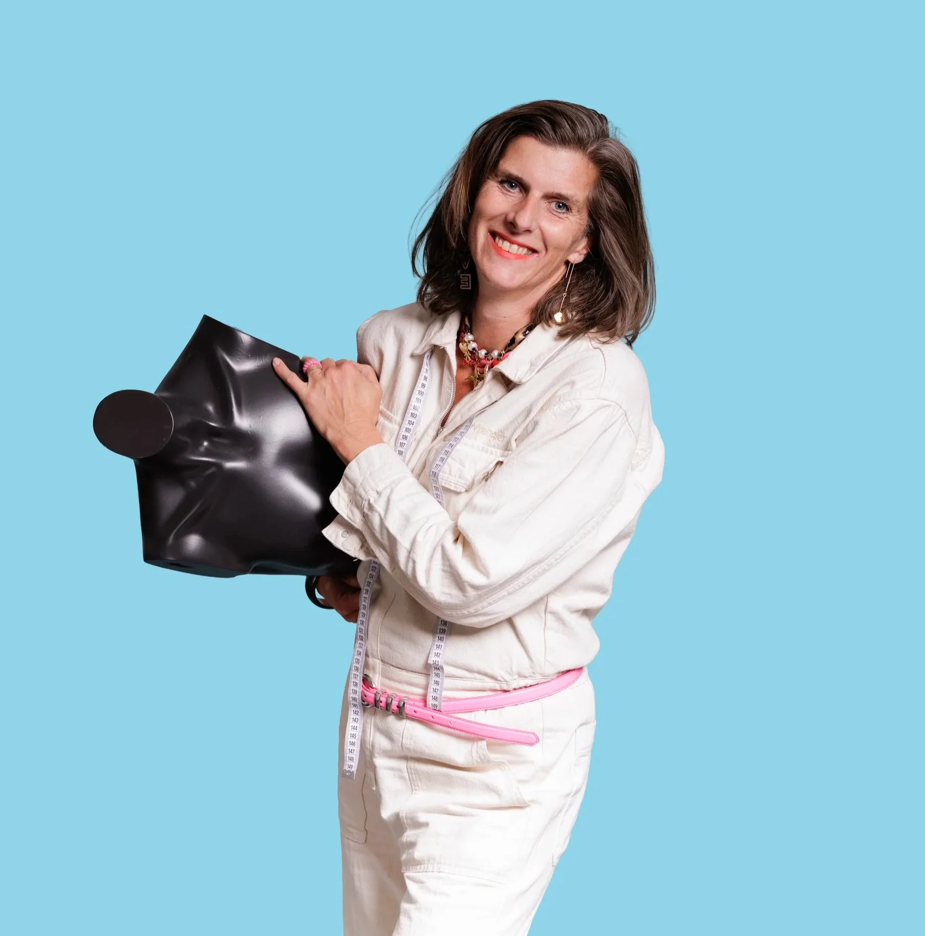 Woman with shoulder-length brown hair smiling, holding a black bust mannequin, wearing a beige jacket, colorful jewelry, and a pink measuring tape around her waist, against a blue background.