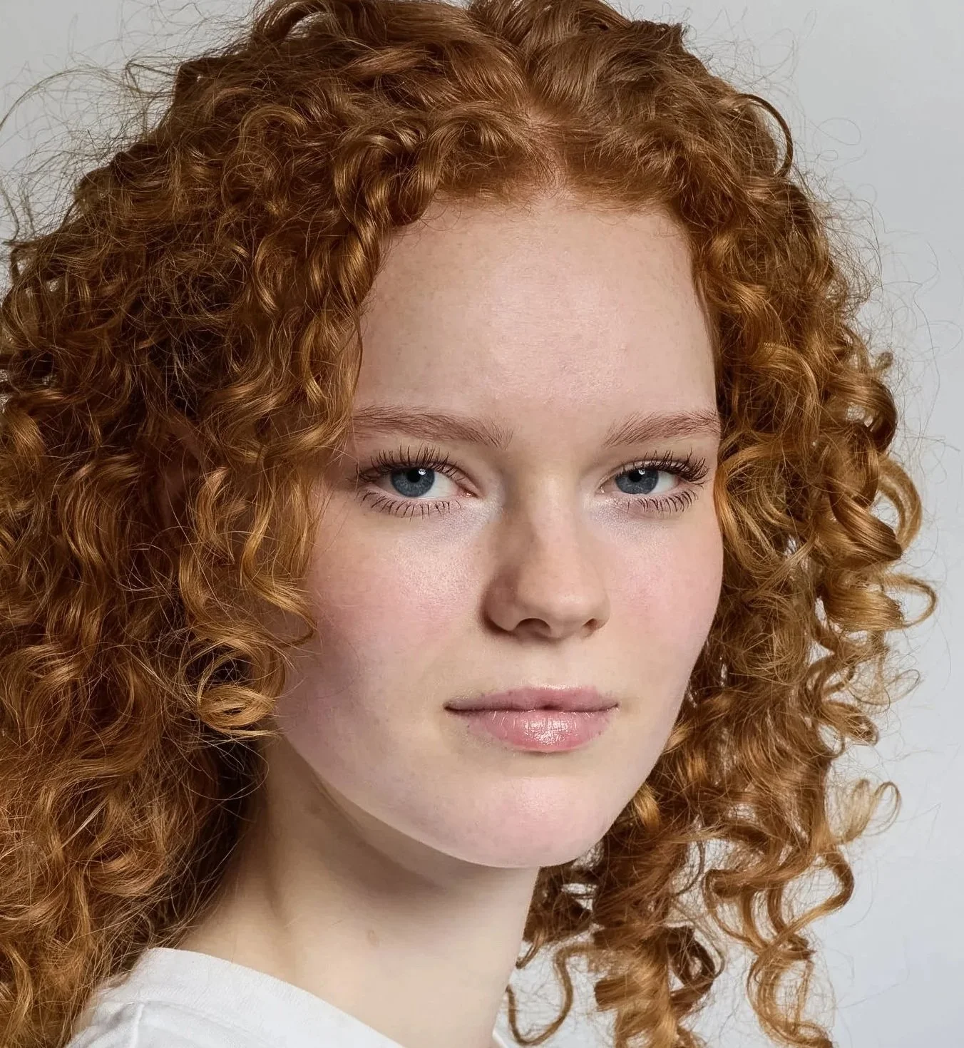 Close-up of a young woman with red curly hair wearing a white top against a plain grey background.