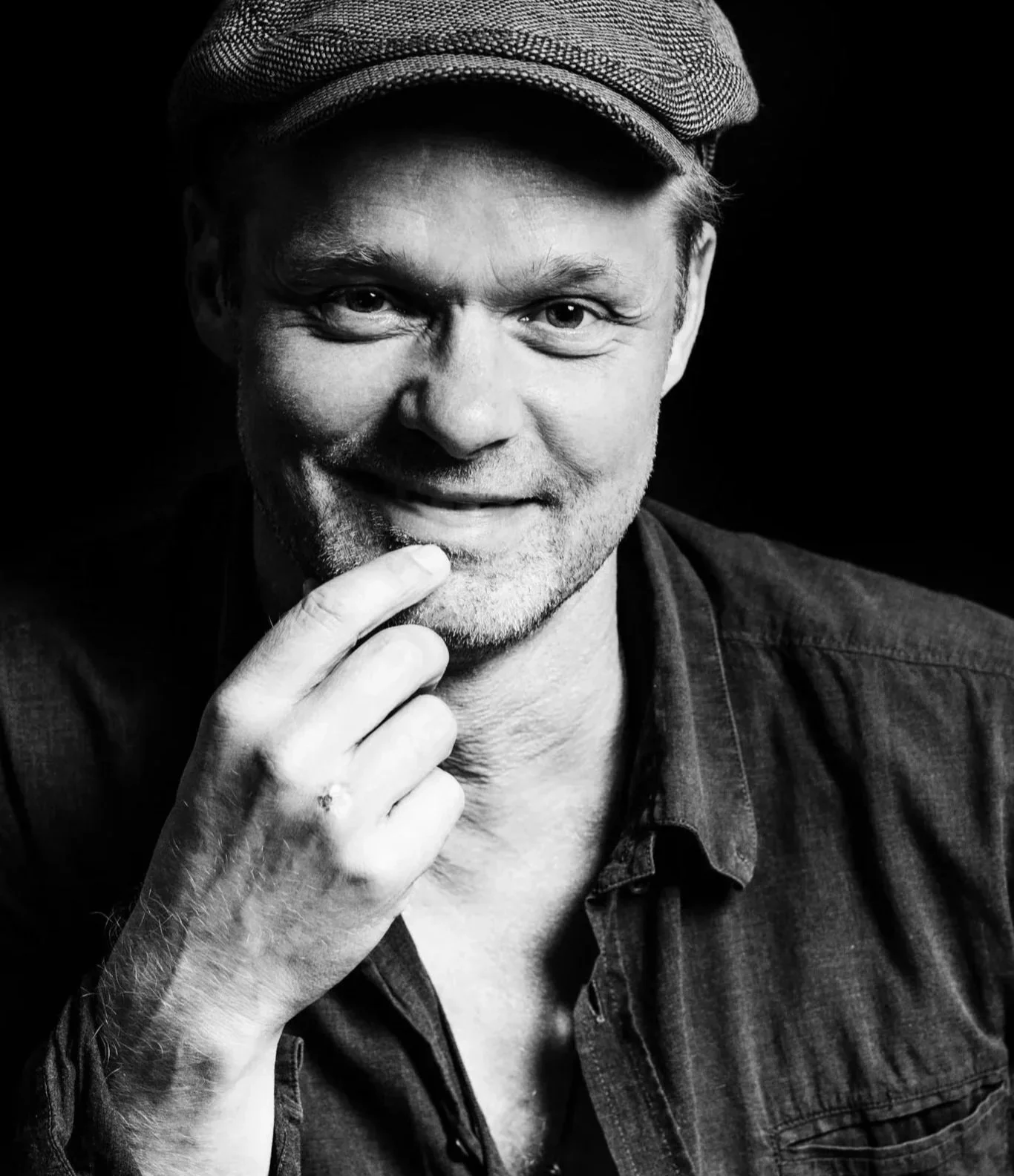 Black and white portrait of a smiling man wearing a flat cap, with his hand resting on his chin.