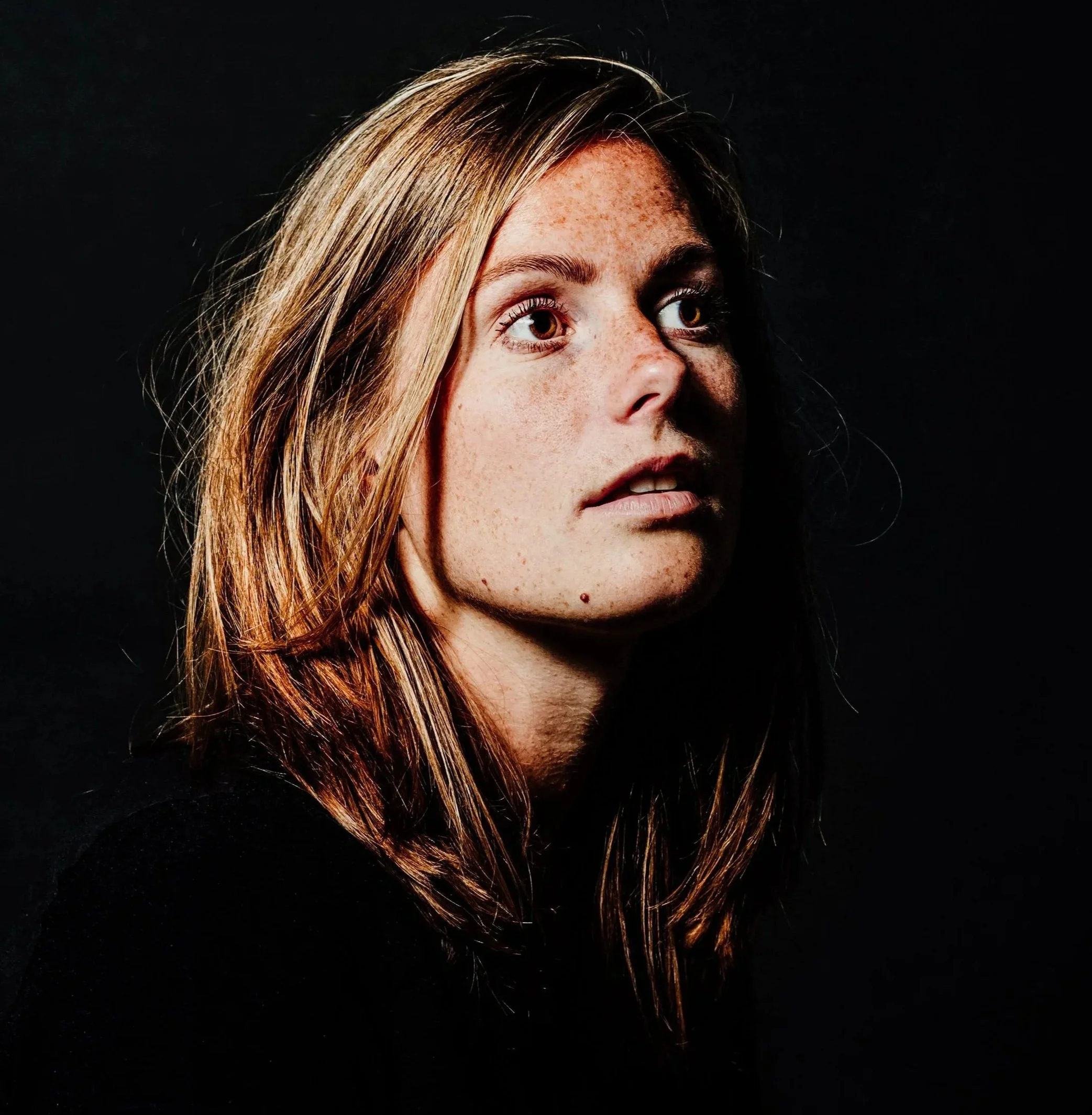 Close-up of a woman's face with freckles, looking to the side, against a black background.