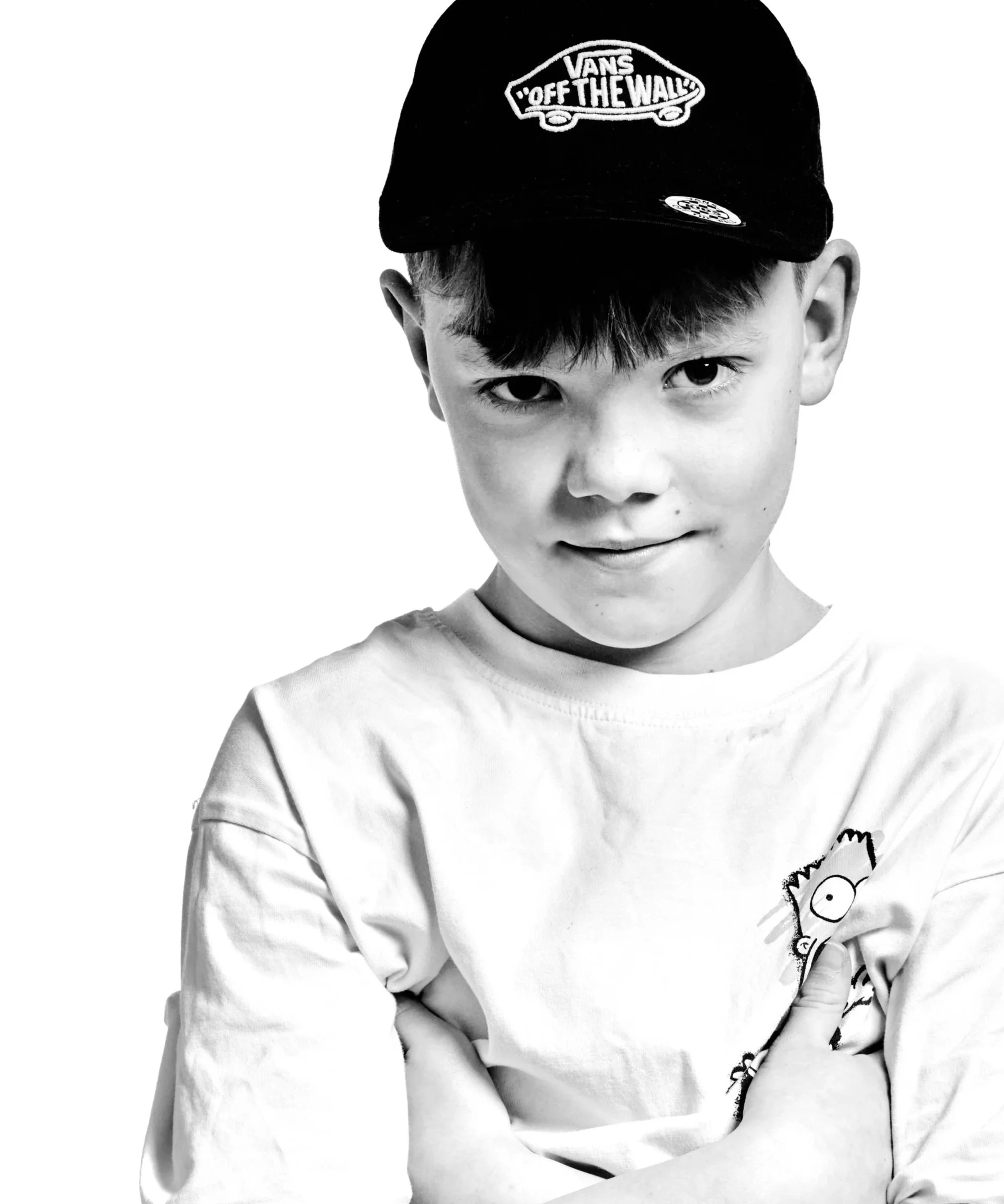 Black and white photo of a young boy wearing a Vans cap and a long-sleeved shirt with a cartoon graphic on the front, standing against a plain background.