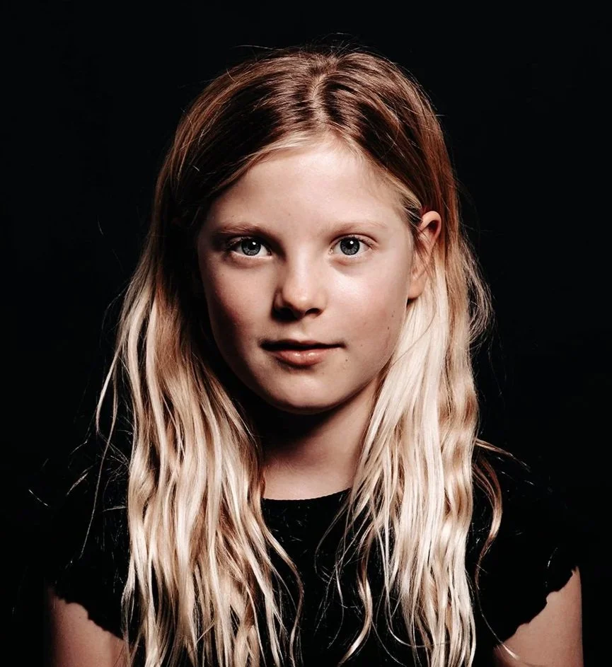Portrait of a young girl with long blonde wavy hair and blue eyes, wearing a black top, against a dark background.