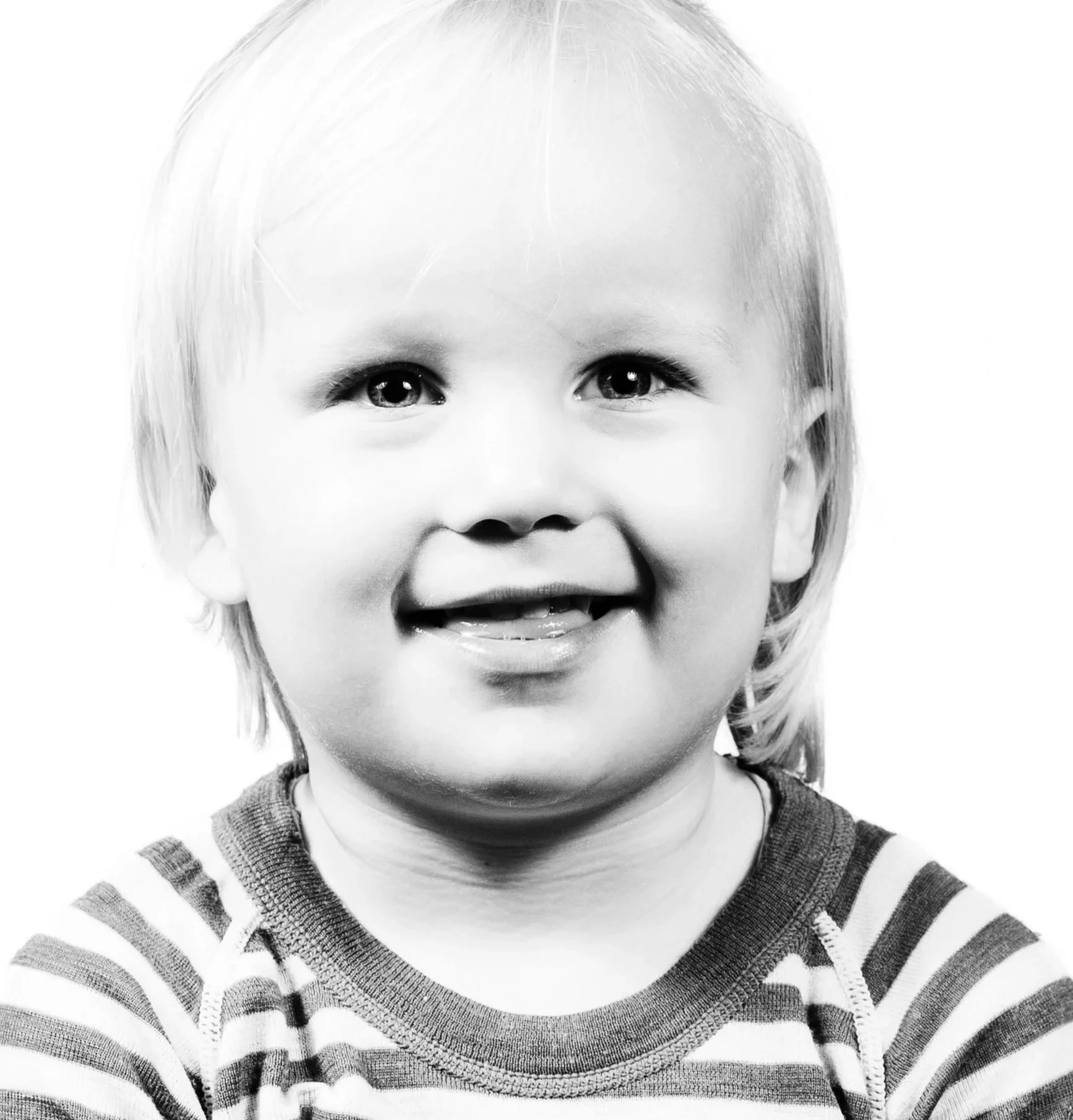 Black and white image of a happy young boy with blond hair, wearing a striped long-sleeved shirt, smiling with his teeth visible, facing forward.