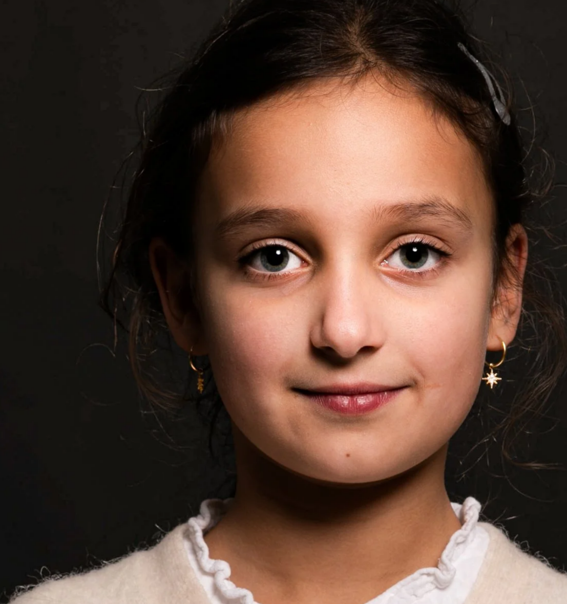 Close-up of a young girl with light complexion, gray eyes, wearing a cream-colored sweater with buttons and star-shaped earrings, against a dark background.