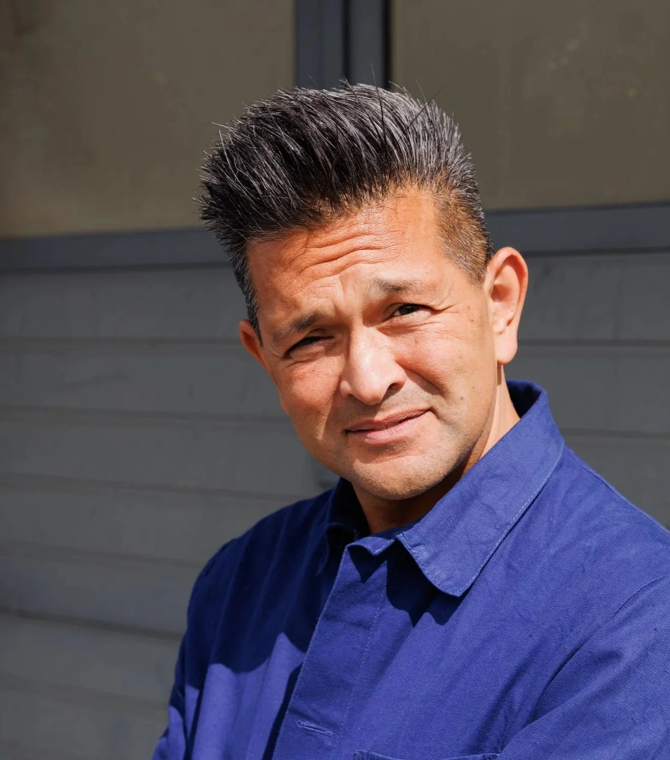 A man with dark, spiky hair and a light complexion wearing a blue collared shirt, standing outdoors in front of a gray building with siding.