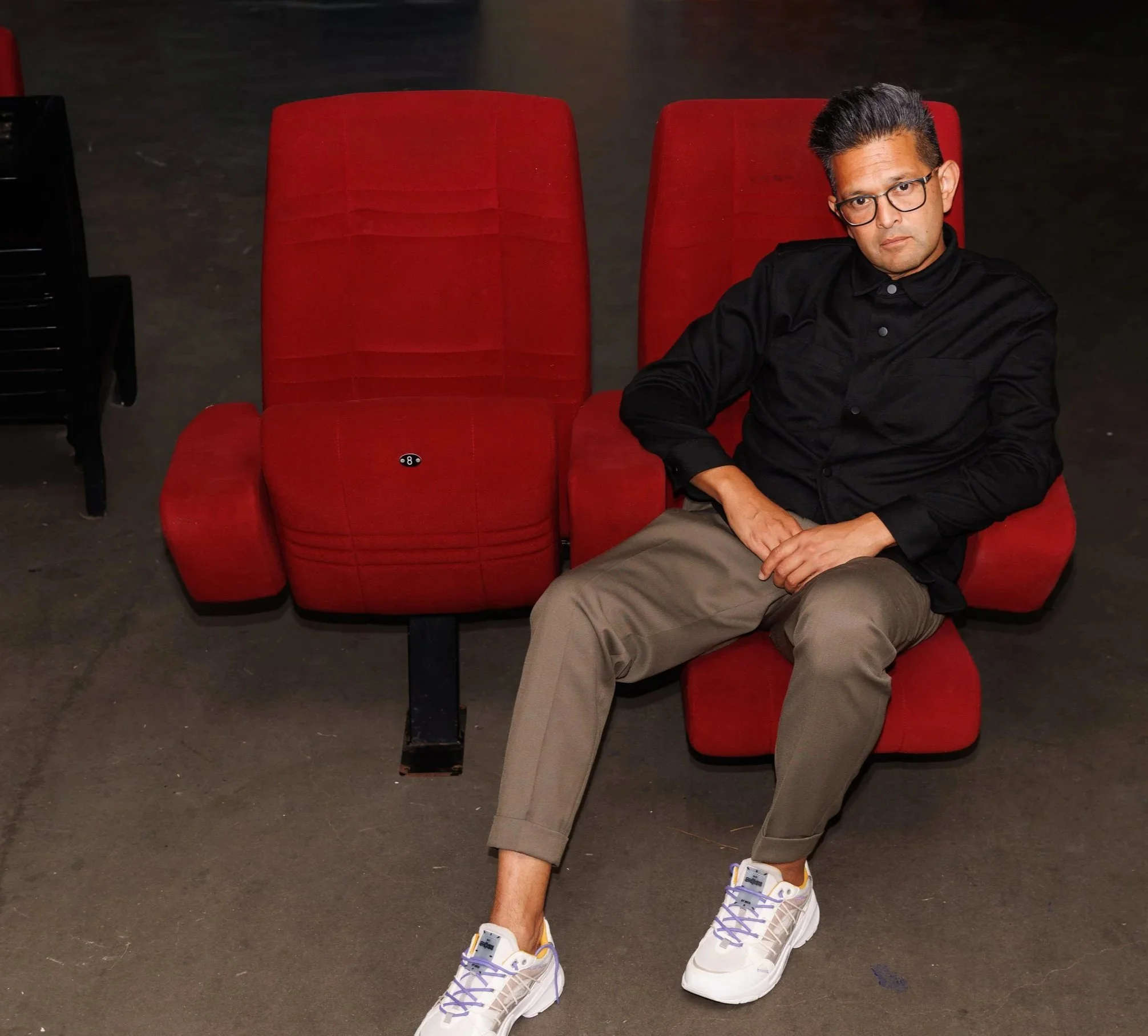 A man with black framed glasses, wearing a black shirt and beige pants, sitting on a red theatre seat with his right arm resting on the armrest, in a dark room with some black chairs and a black object on the left side of the image.