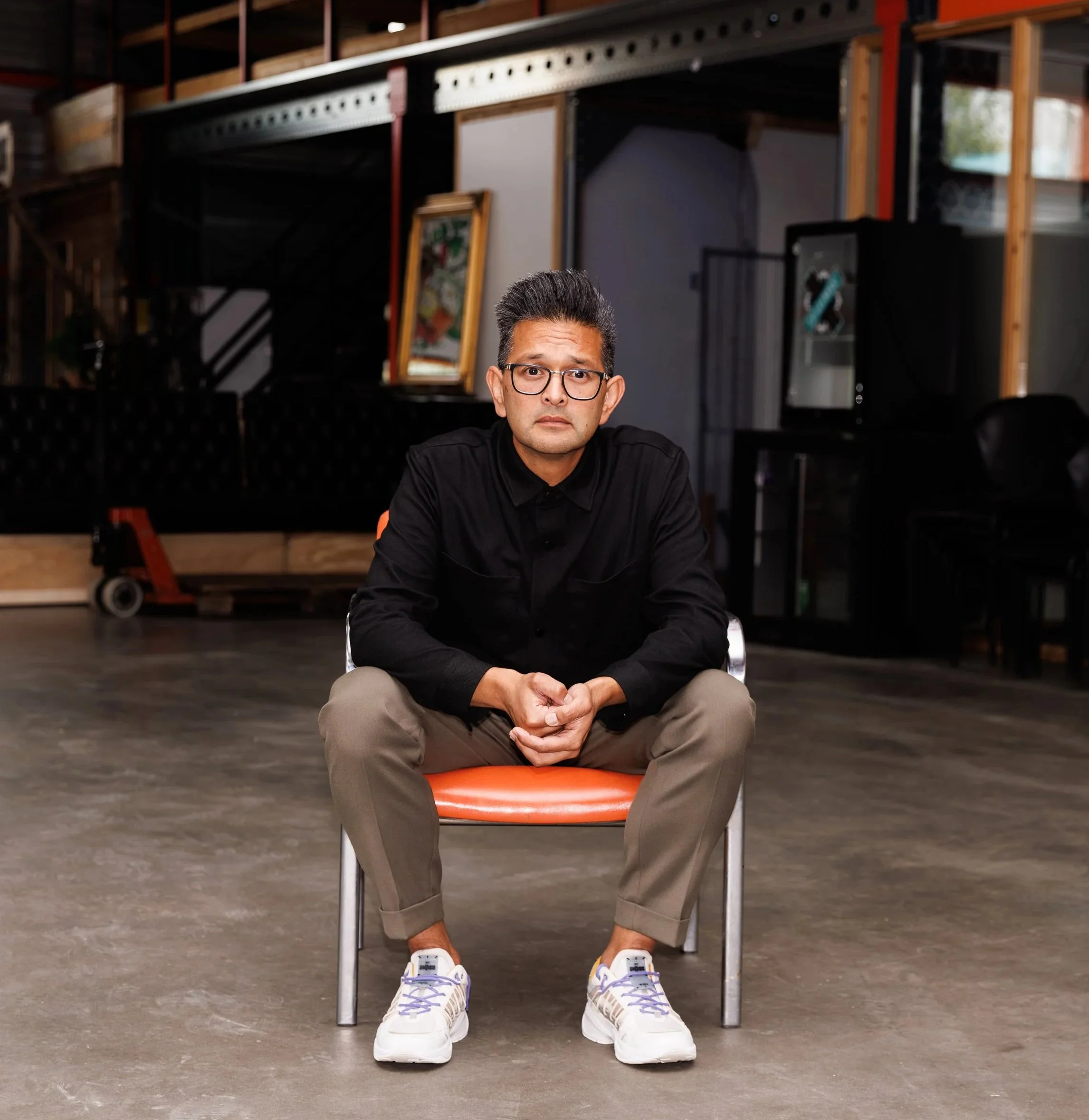 A man sitting on an orange chair in an industrial-style space, wearing glasses, a black shirt, and beige pants, with sneakers. The background includes shelving, paintings, black equipment, a dolly, and a window.