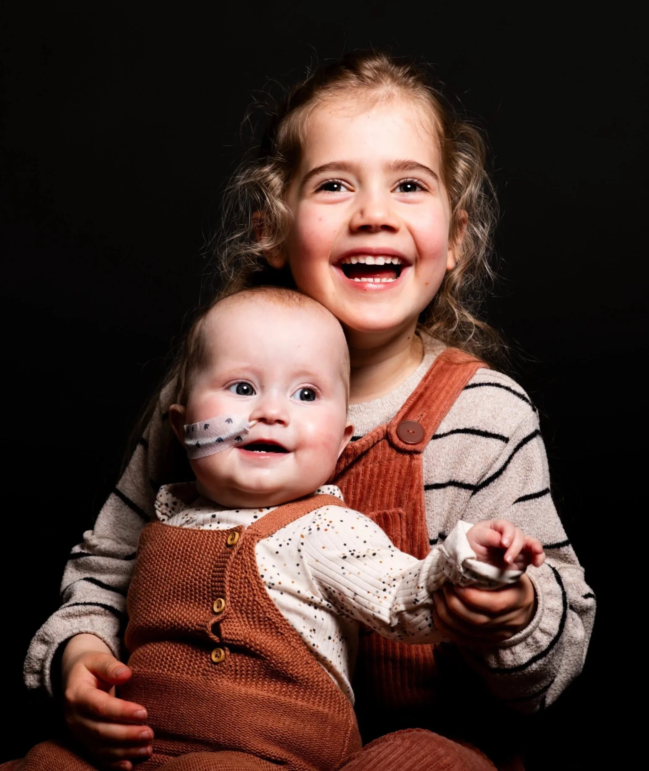 Twee lachende jonge meisjes, een met bruin haar en een met blond haar, poseren tegen een zwarte achtergrond.