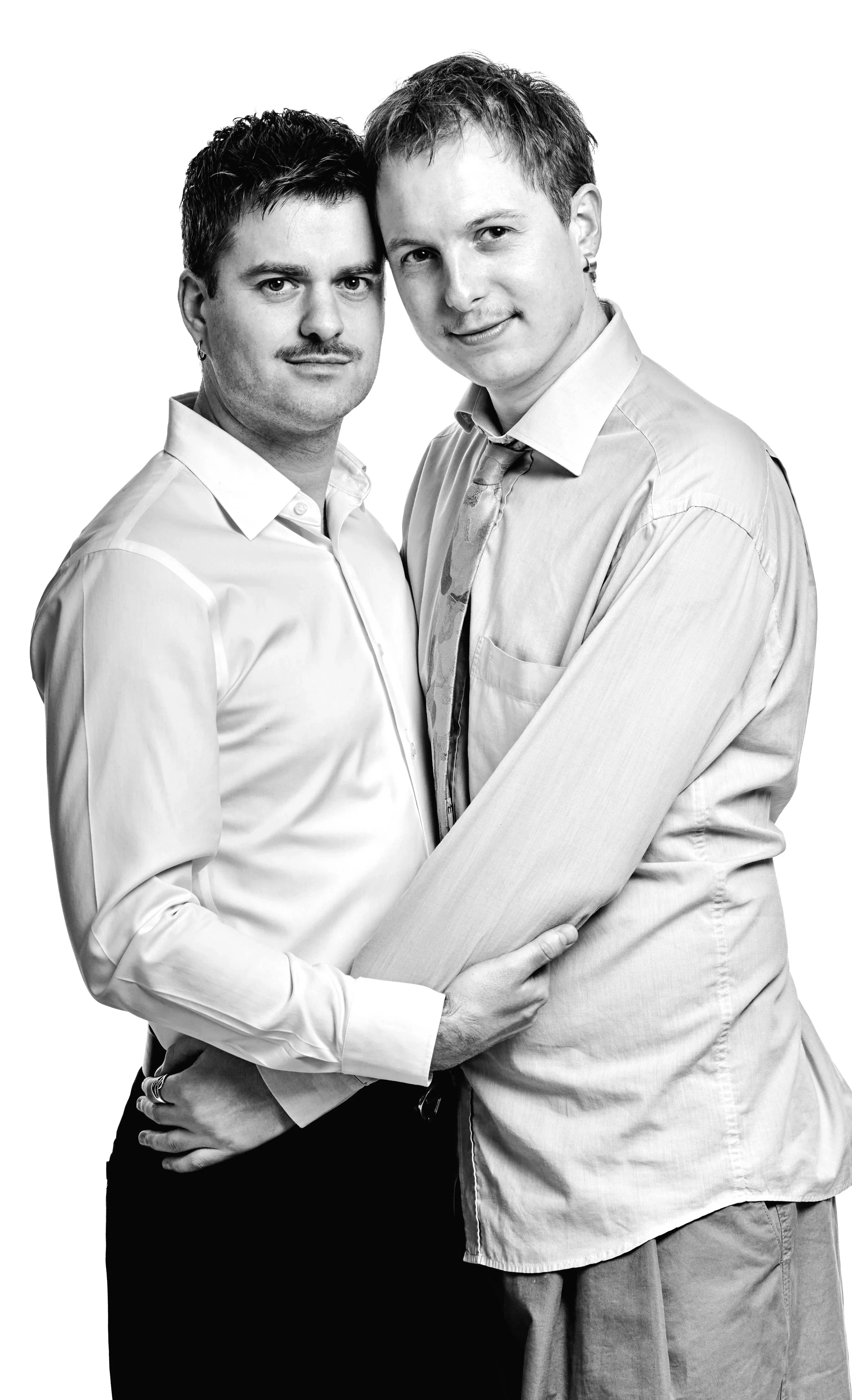 Two men standing close together, holding each other with their arms around each other's waists, smiling at the camera, in grayscale.